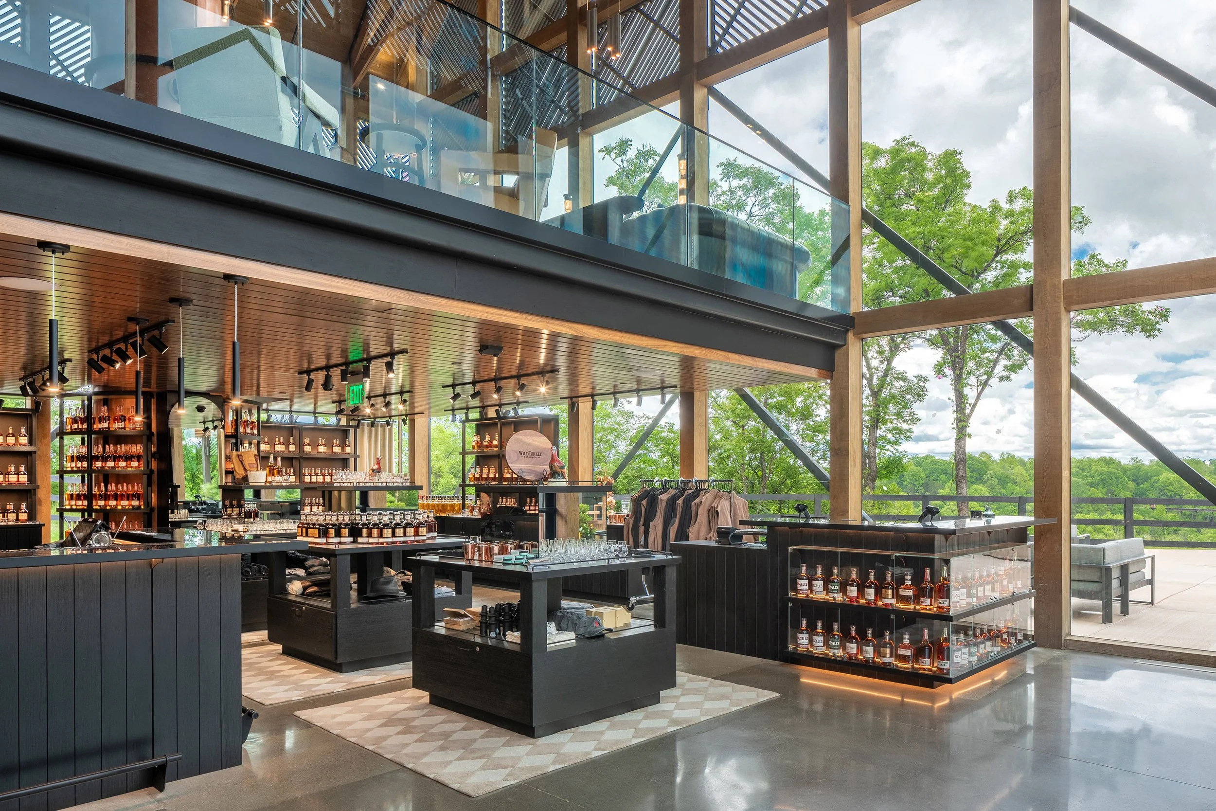 Modern retail store with large glass windows, wooden beams, and shelves displaying bourbon bottles and merchandise. Photography displays highlight clothing items, while trees and a cloudy sky are visible outside the visitor center.