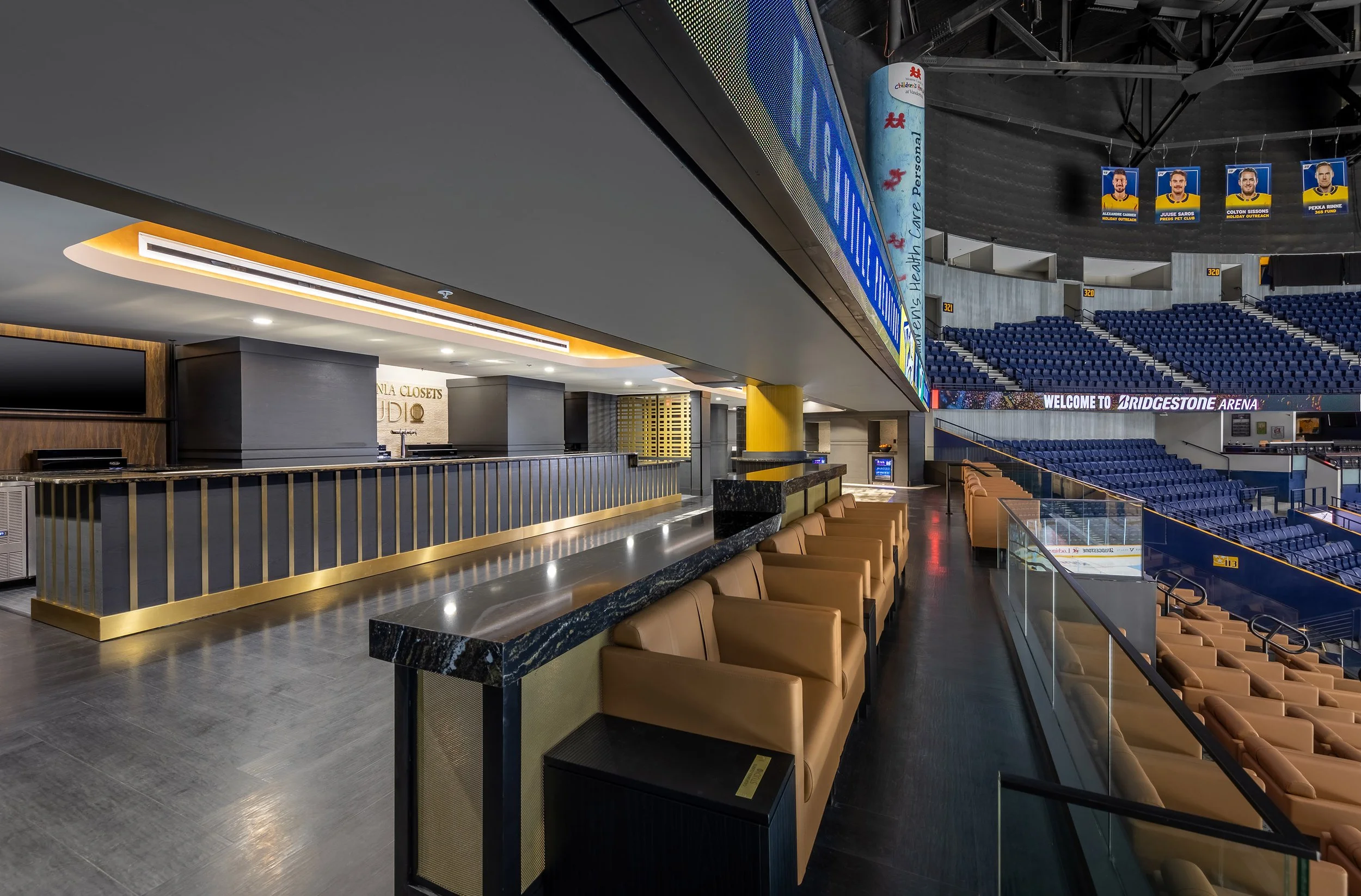 Luxury Club suite with tan armchairs and black marble countertops overlooks an indoor sports arena. The suite features a sleek bar area, TVs, and a clear view of the hockey rink and scoreboard in the Predators' Arena.