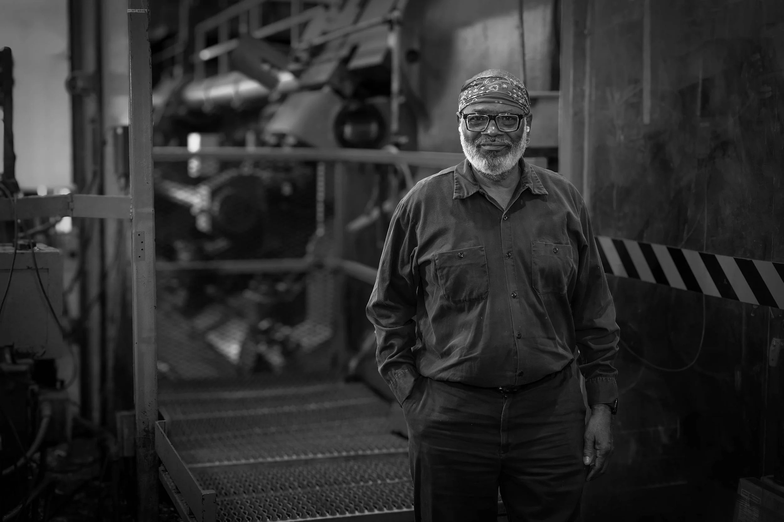 Older man with a beard, wearing glasses and a bandana, smiles in a black and white industrial setting.