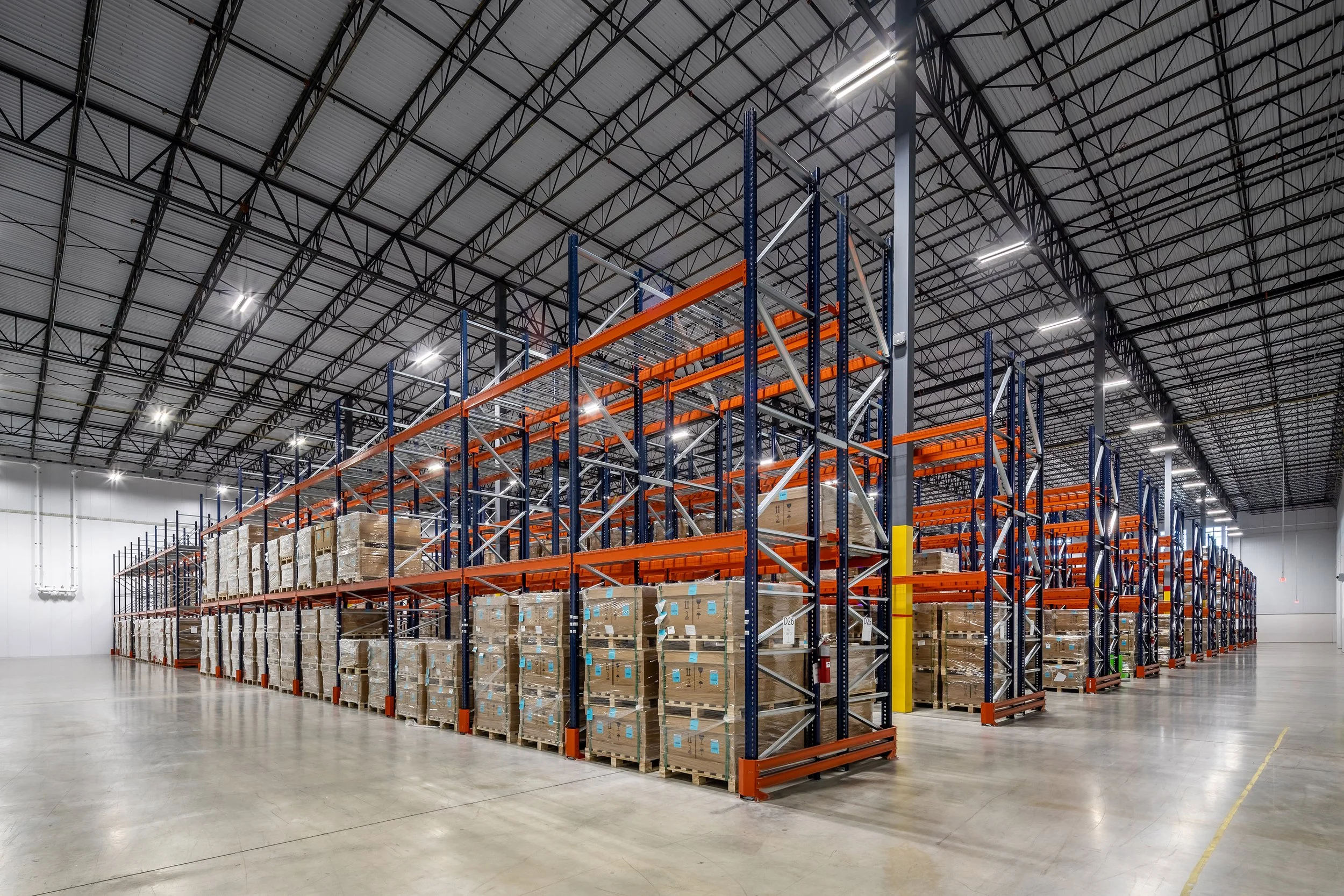 Spacious industrial warehouse interior in Texas featuring tall metal racks stacked with cardboard boxes on pallets, under high ceilings with exposed beams, bright lighting, and a polished concrete floor.
