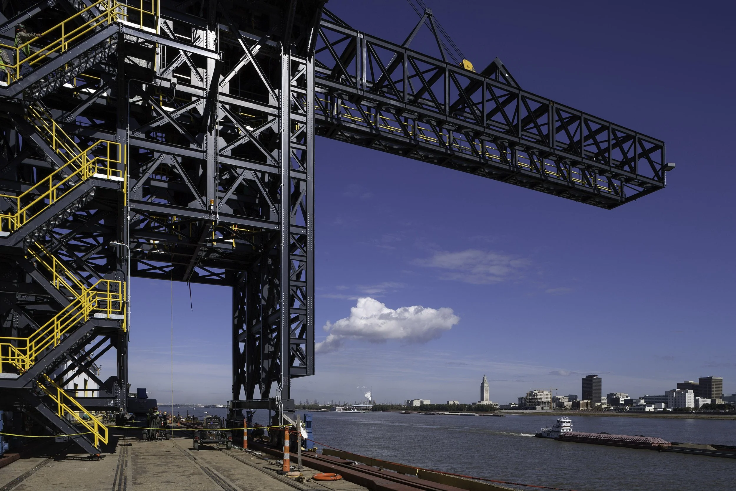 Huge industrial crane with yellow stairs beside a river, city skyline and blue sky in the background.