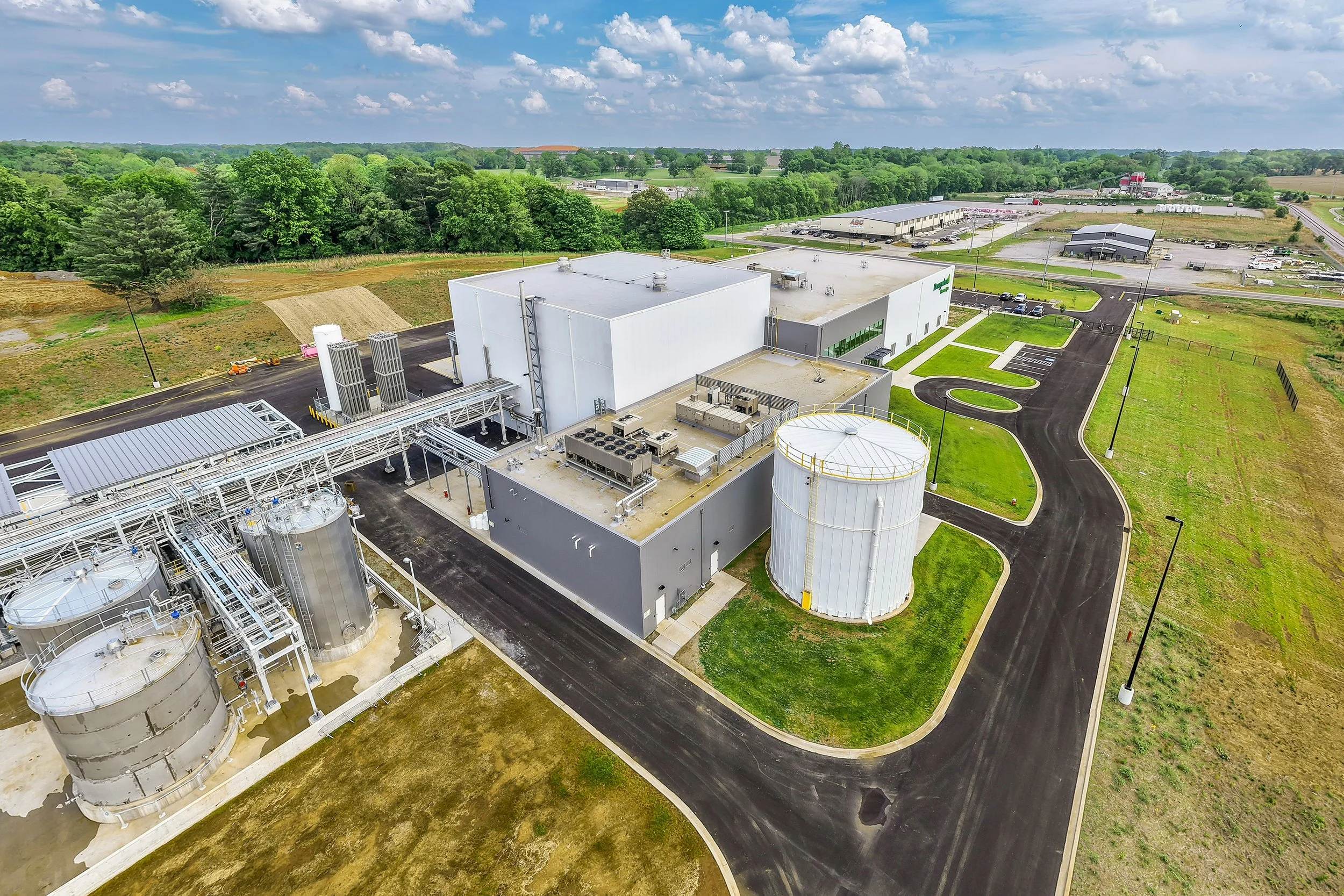 Aerial photography captures an industrial facility with several large white tanks, gray and white buildings, paved roads, and grassy areas under a partly cloudy sky. Trees and additional structures enhance the architectural landscape in the backgroun