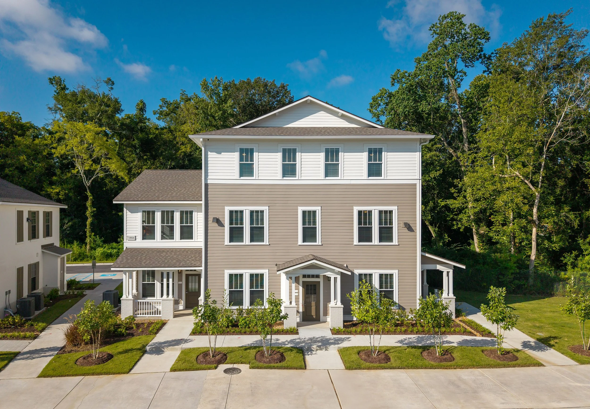A modern three-story multi family house with gray and white siding, large windows, and a front porch, surrounded by young trees and landscaped lawns, with a forested area in the background under a blue sky.