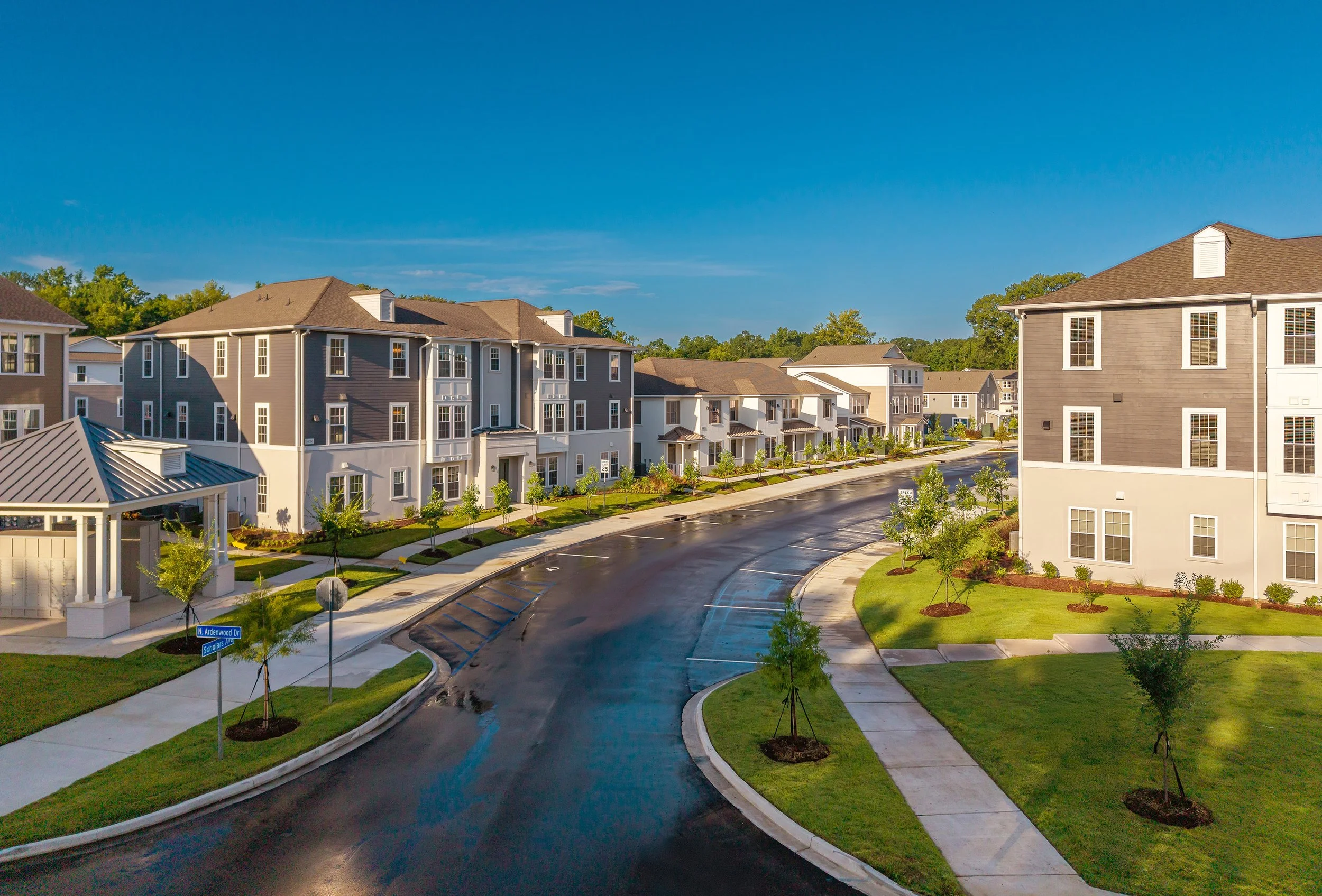 A freshly paved road curves through a modern residential neighborhood with multi-story apartment buildings, green lawns, and neatly landscaped trees under a clear blue sky.