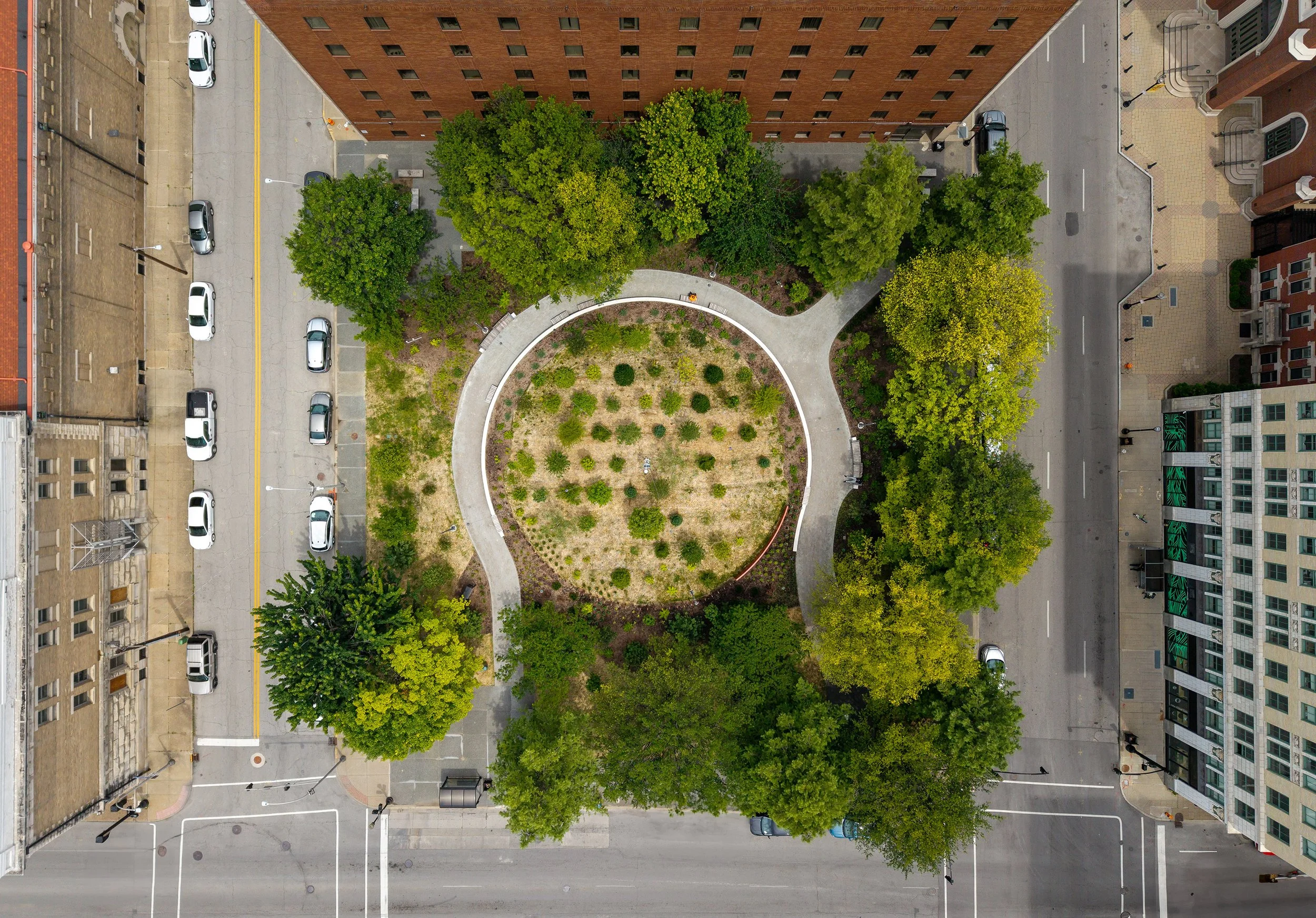 Aerial view of an urban park with circular landscape design, bordered by trees and paths, surrounded by city streets and parked cars; buildings line the edges of the block.