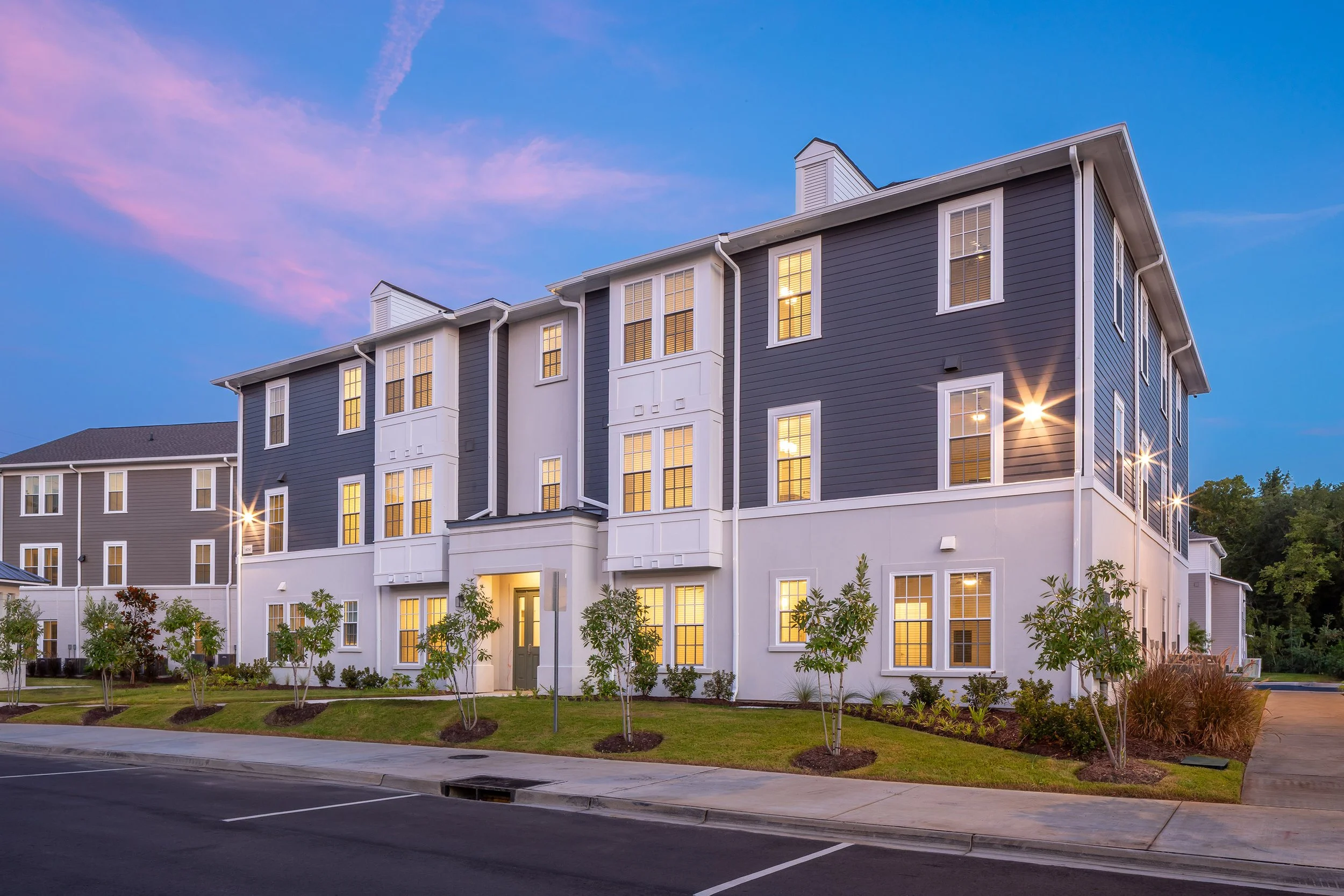 Modern three-story apartment building with gray and white exterior, lit windows, landscaping with small trees, and a parking lot in front, photographed at dusk with a blue and pink sky.