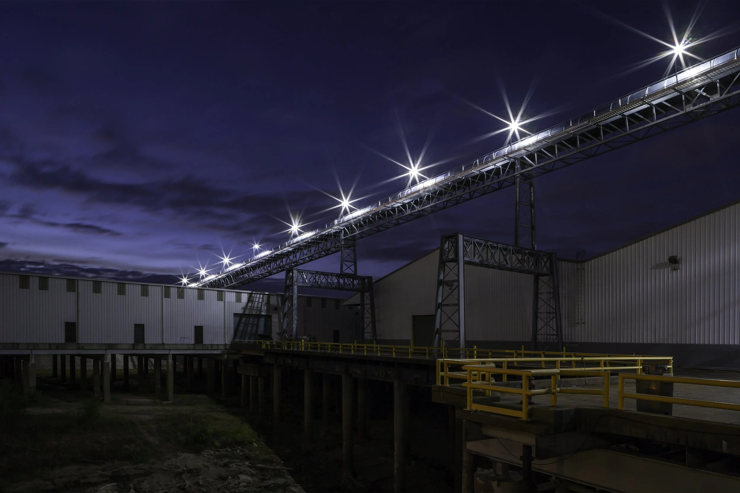 Industrial building at night with bright lights on an elevated conveyor against a dark, cloudy sky.
