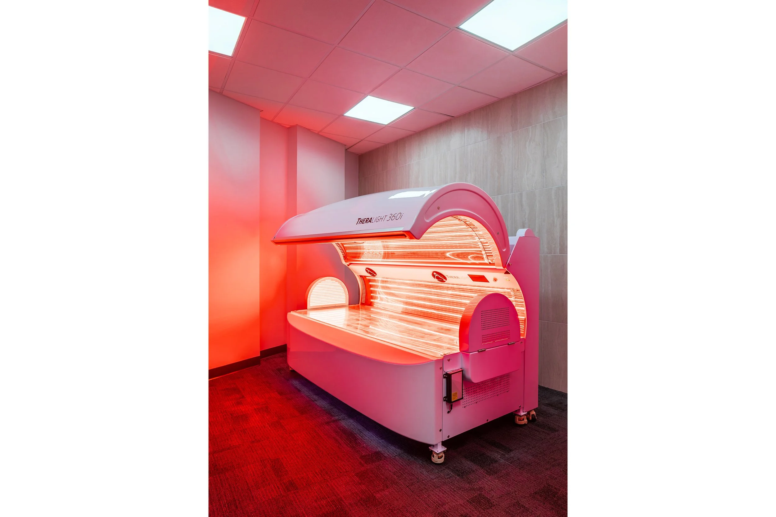 A therapy bed glows with bright red light in a small recovery room with gray walls, red carpet, and fluorescent ceiling lights. The bed is open and unoccupied, revealing its illuminated interior.
