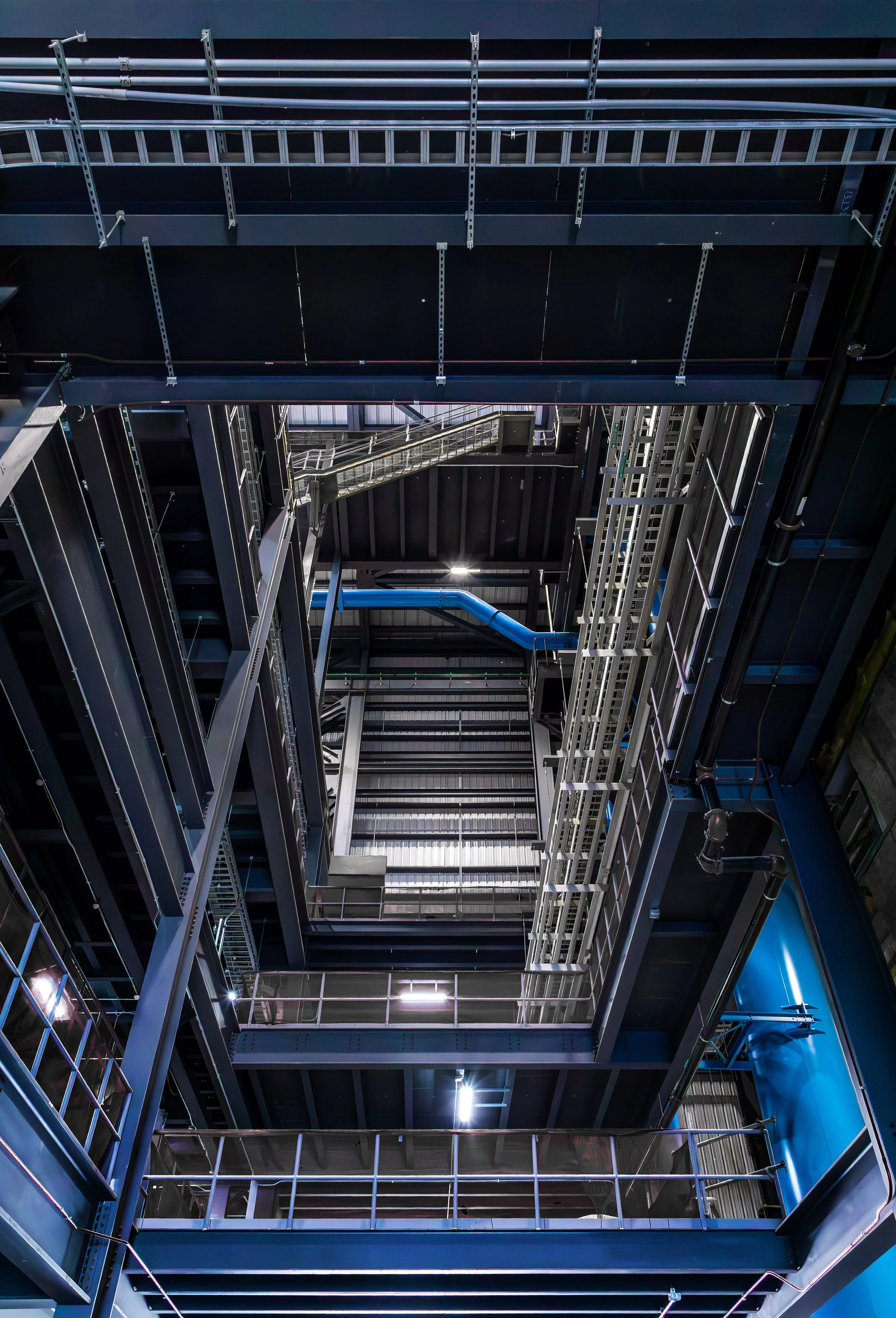 View looking up at multiple levels of architectural metal walkways, platforms, and structural beams inside an industrial building, with geometric lines and dramatic lighting creating a sense of depth and complexity.