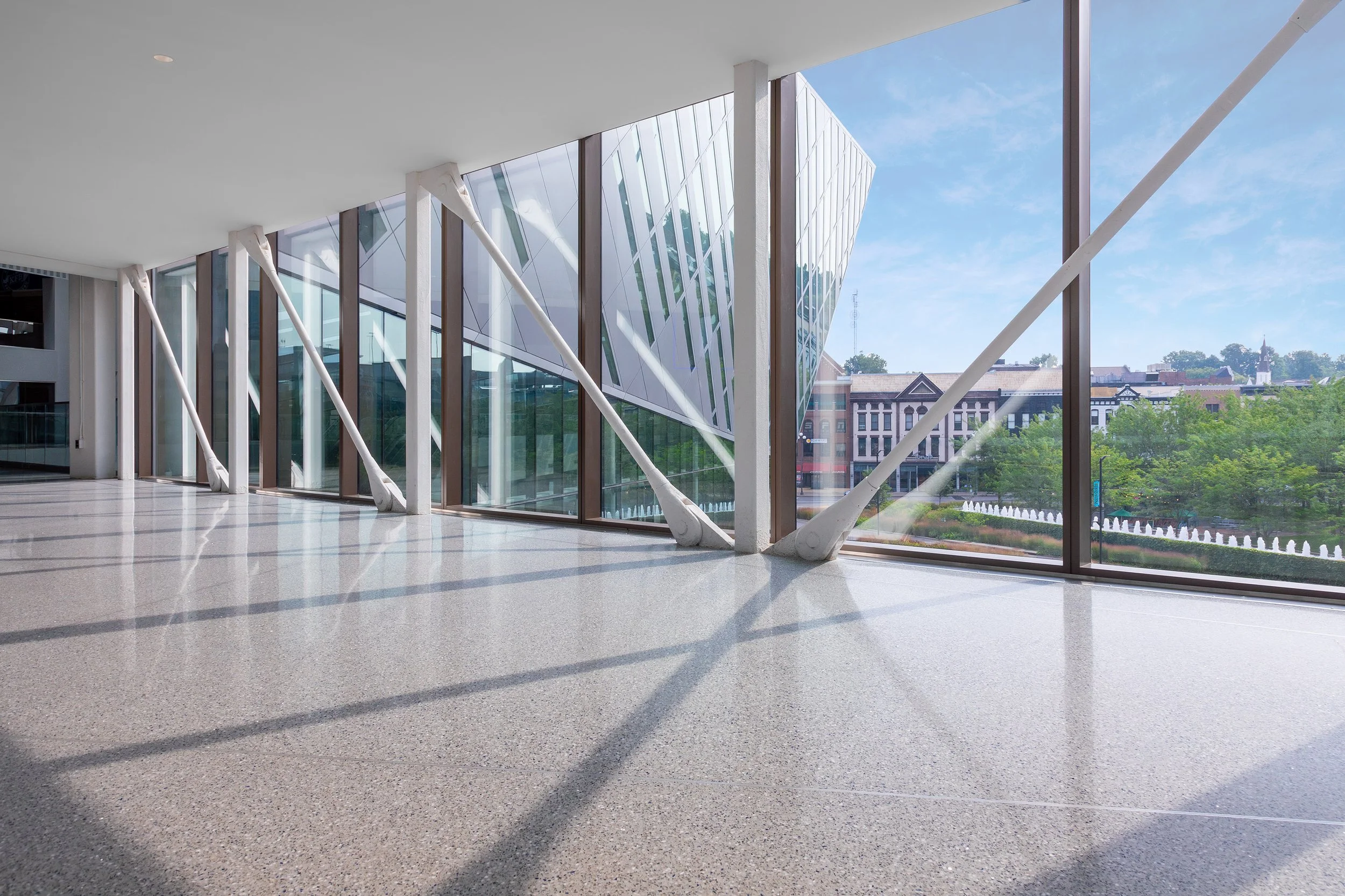 Sunny hallway with floor-to-ceiling windows, diagonal beams, and a view of modern and historic buildings from the pedestrian walkway leading to Rupp Arena.