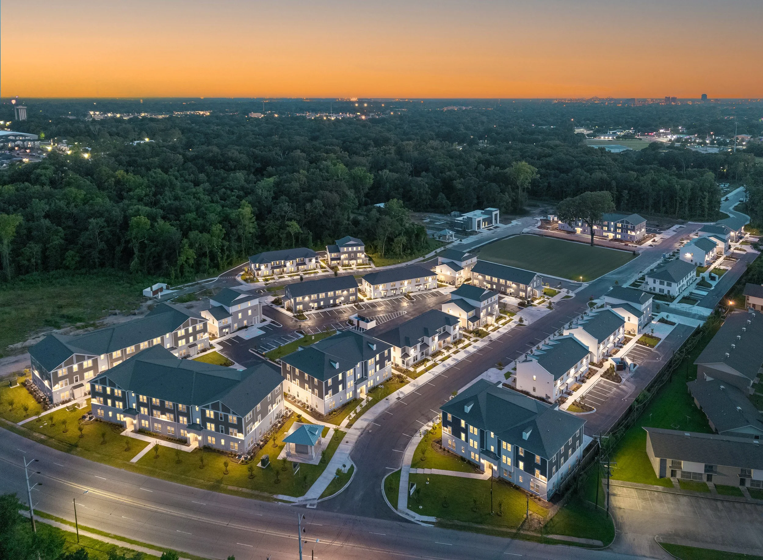 Aerial view of a modern residential complex at dusk, with illuminated buildings, curved roads, and surrounding greenery. The sky is orange, and city lights are visible in the distant background.