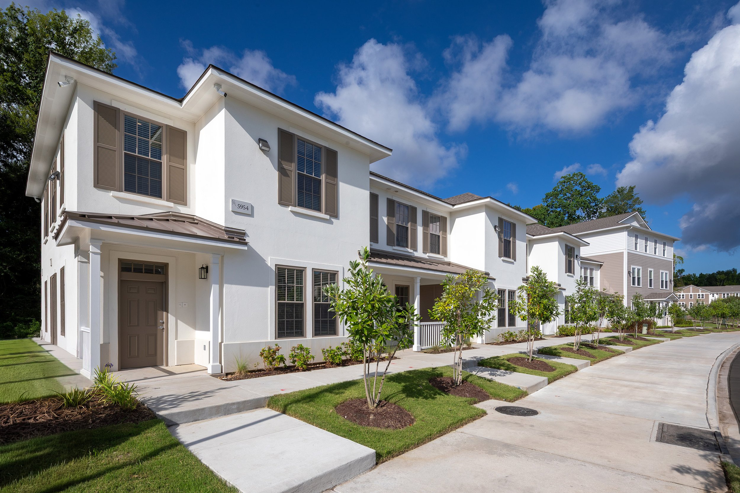 Row of modern, two-story white townhouses with manicured lawns, young trees, and a clean sidewalk under a bright blue sky with scattered clouds.