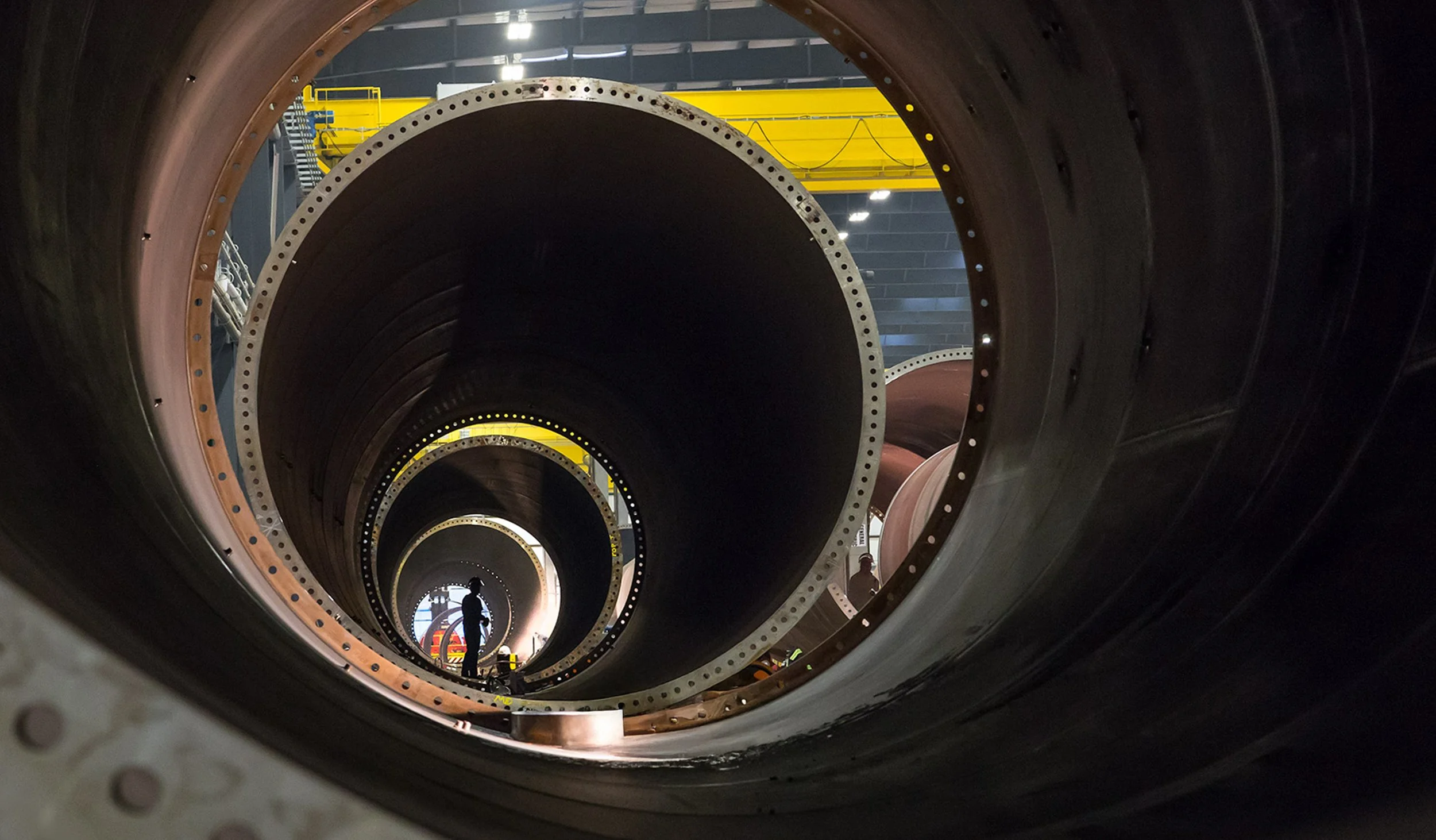 View of wind turbine towers from inside a factory or power plant with a worker standing in the distance, surrounded by machinery and yellow overhead equipment.