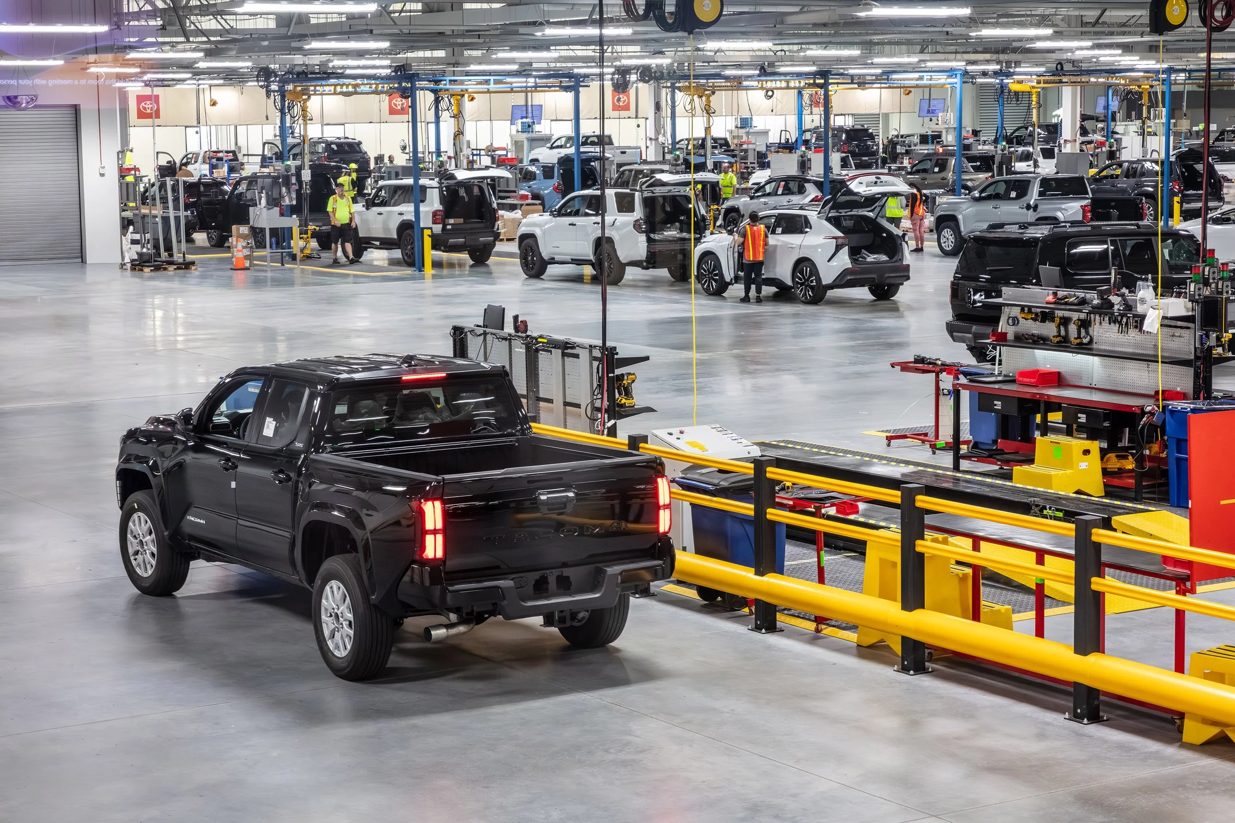 A black pickup truck drives through a brightly lit automotive factory where several vehicles are being assembled by workers in safety vests among equipment and assembly stations.