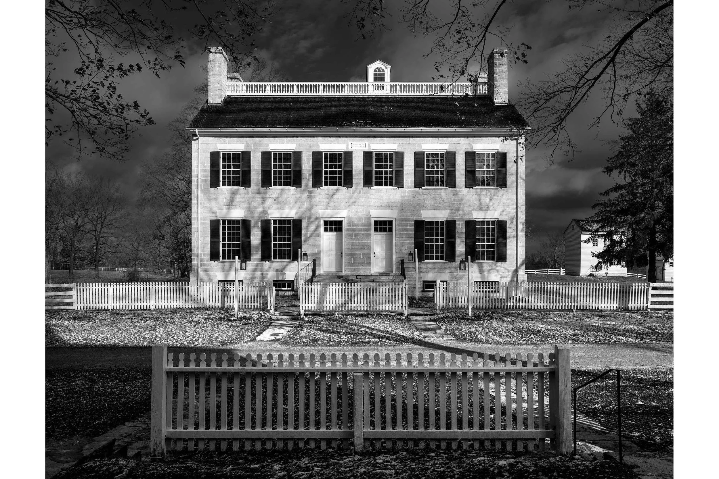 Black-and-white photo of a large Shaker building with columns, shutters, and a white picket fence in a bare winter landscape.