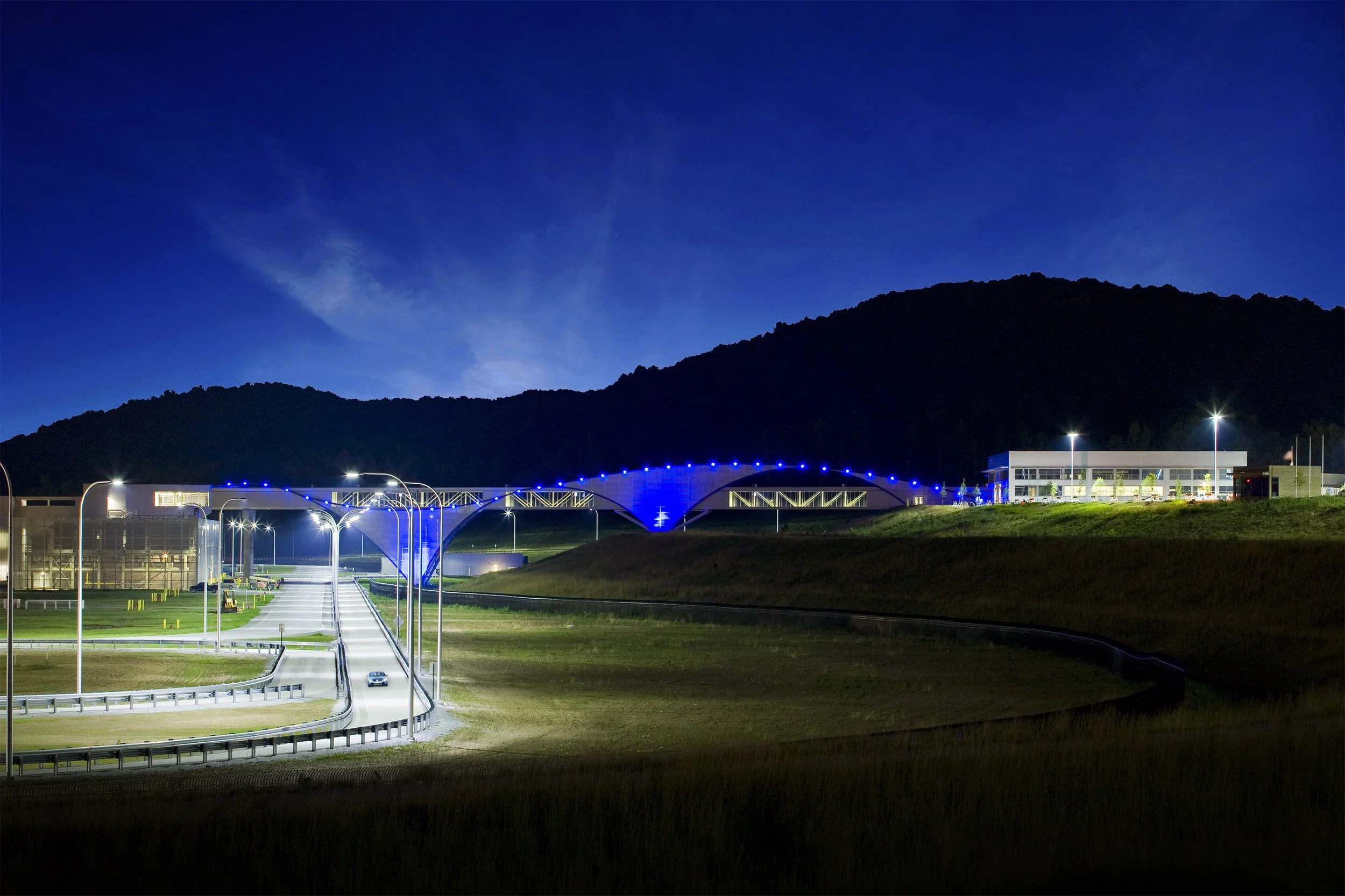 A test track leads from a blue pedestrian bridge at night, with hills, buildings, and a Volkswagen automotive assembly in the background.