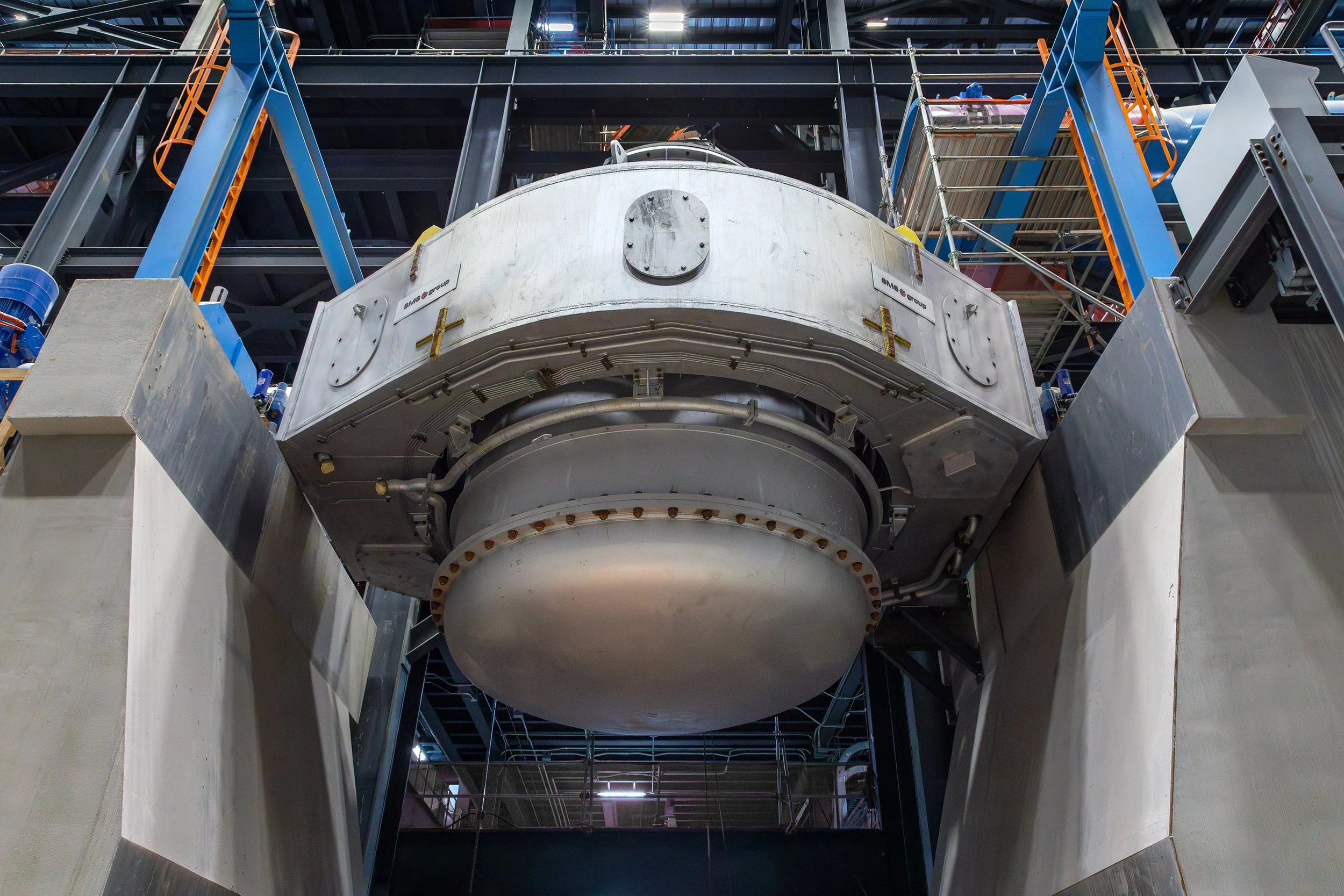 A large, metallic, circular melting vessel is suspended by blue support structures inside an industrial facility, showcasing striking architectural elements with beams and platforms visible in the background.