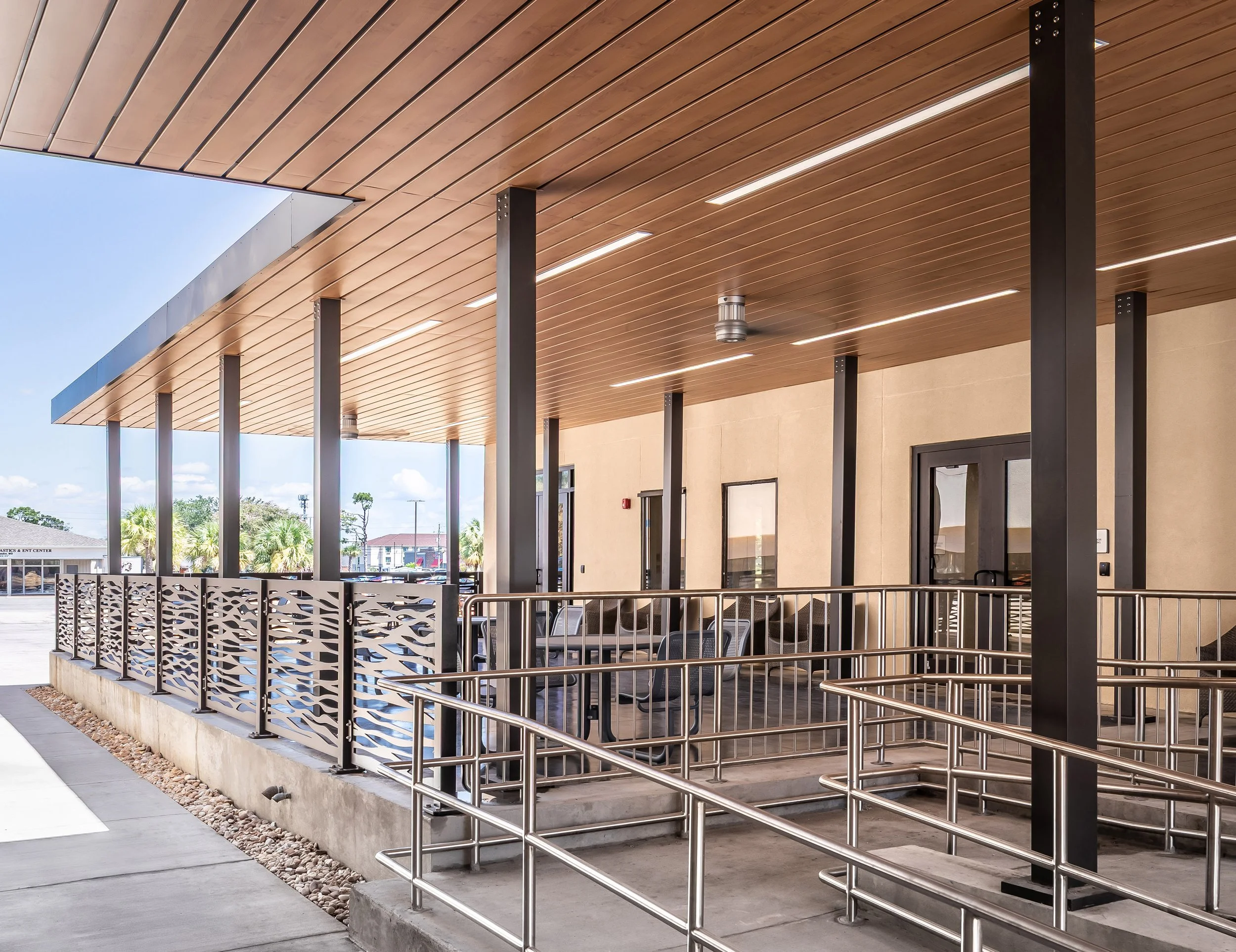 A modern building entrance with a wooden ceiling, metal columns, decorative railing, glass doors, and a wheelchair-accessible ramp creates an inviting outdoor setting under a clear blue sky—perfect for photography.