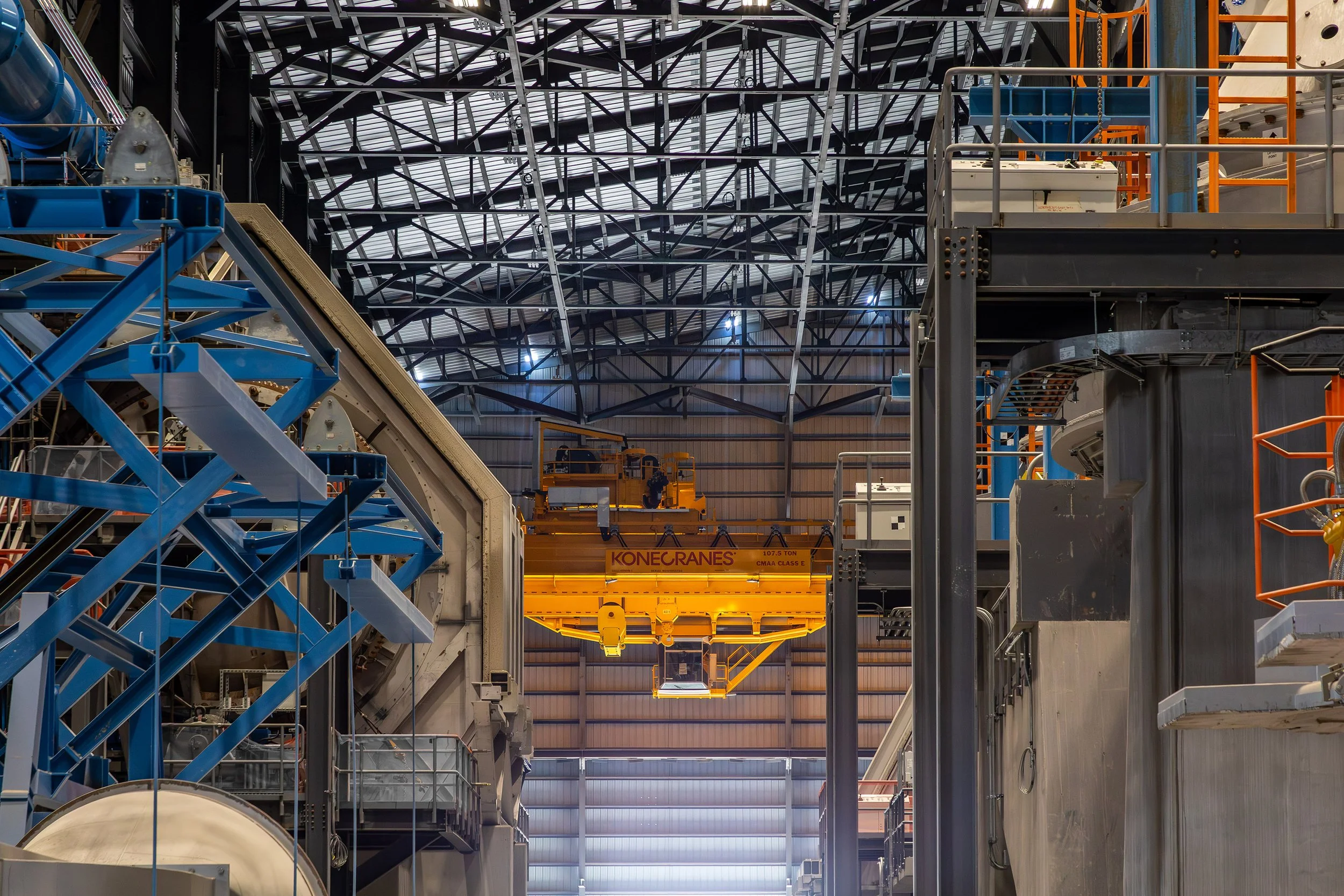 An industrial facility interior with a large yellow Konecranes overhead crane suspended among architectural metal structures, beams, and machinery under a high, metal truss roof.