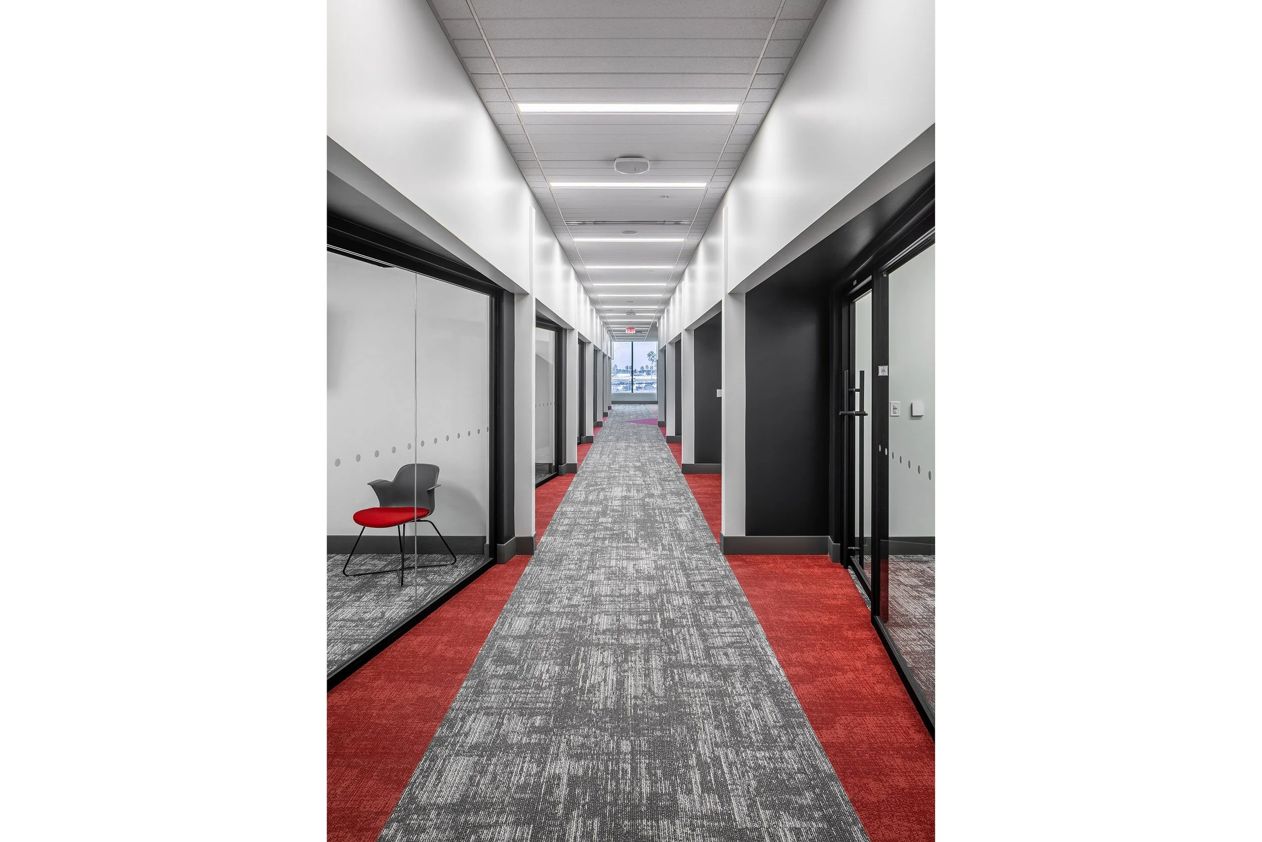 A modern hallway with gray carpet, red accents, glass office doors on both sides, and a single black and red chair visible in one of the offices; ceiling lights run along the corridor.