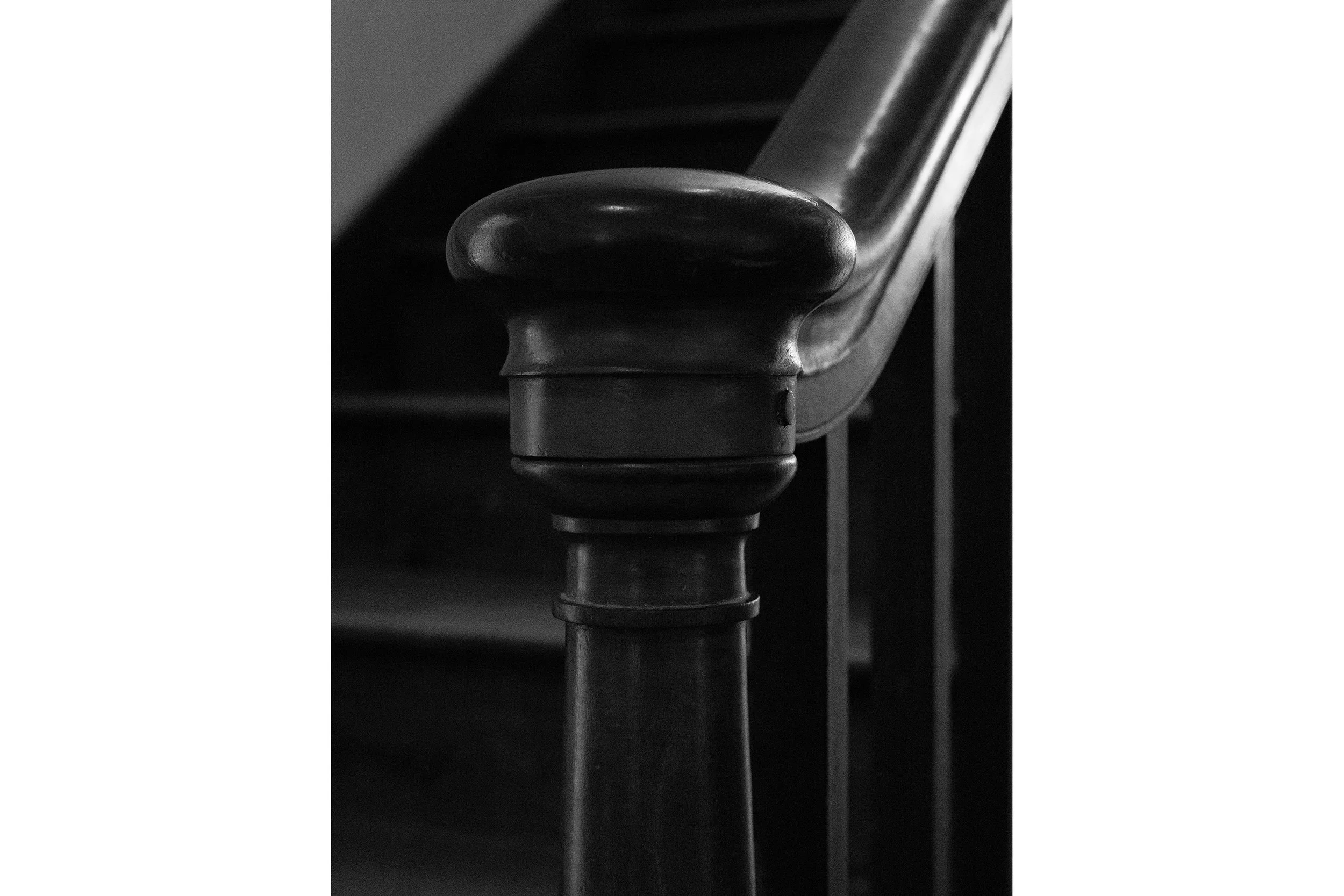 Close-up of a Shaker-style wooden staircase banister in black and white, with blurred stairs fading into the background.