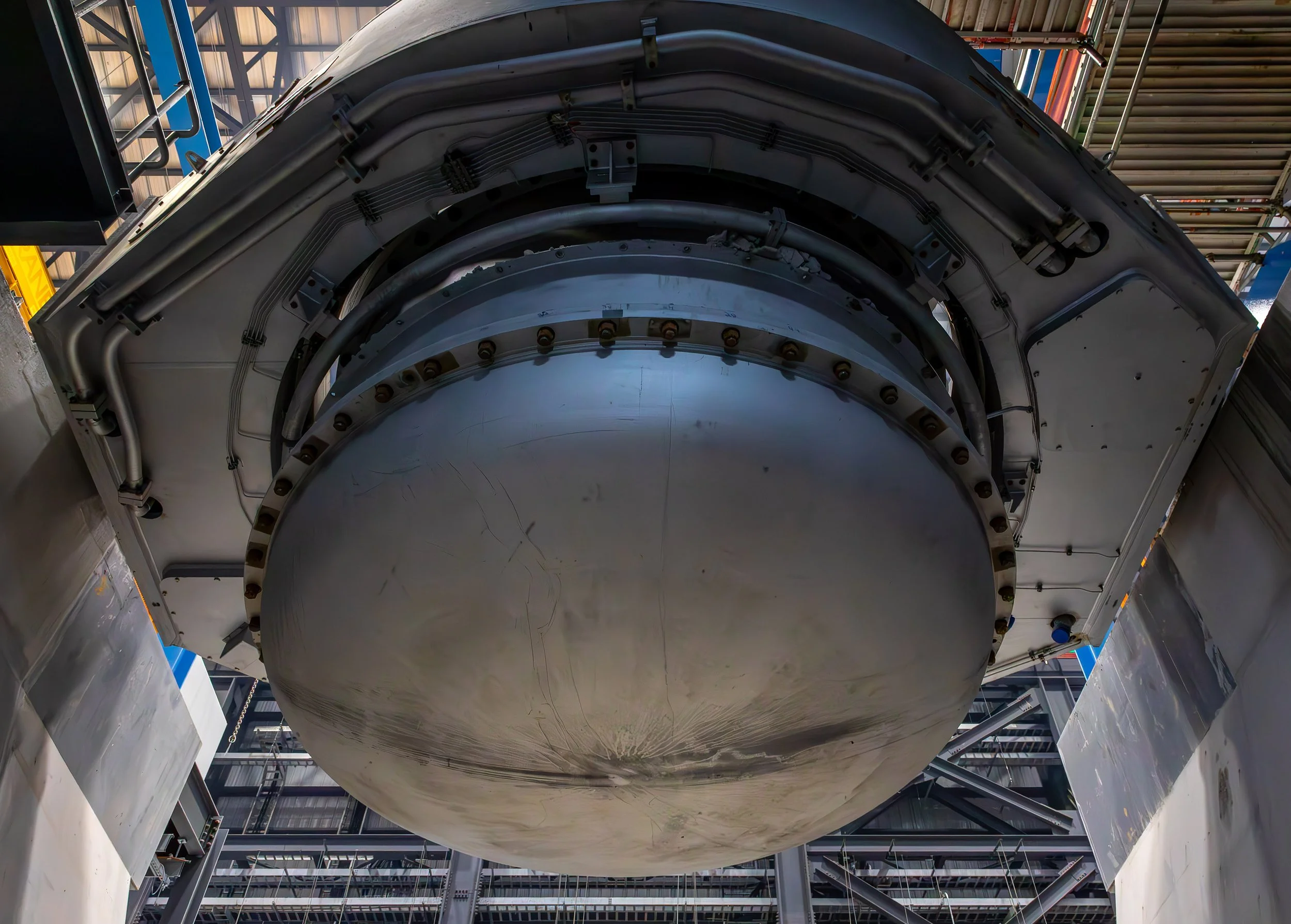 Close-up photography of a large, metallic, circular structure—possibly a melter—with cables and bolts lining the edge, set in an industrial environment filled with beams and pipes overhead.