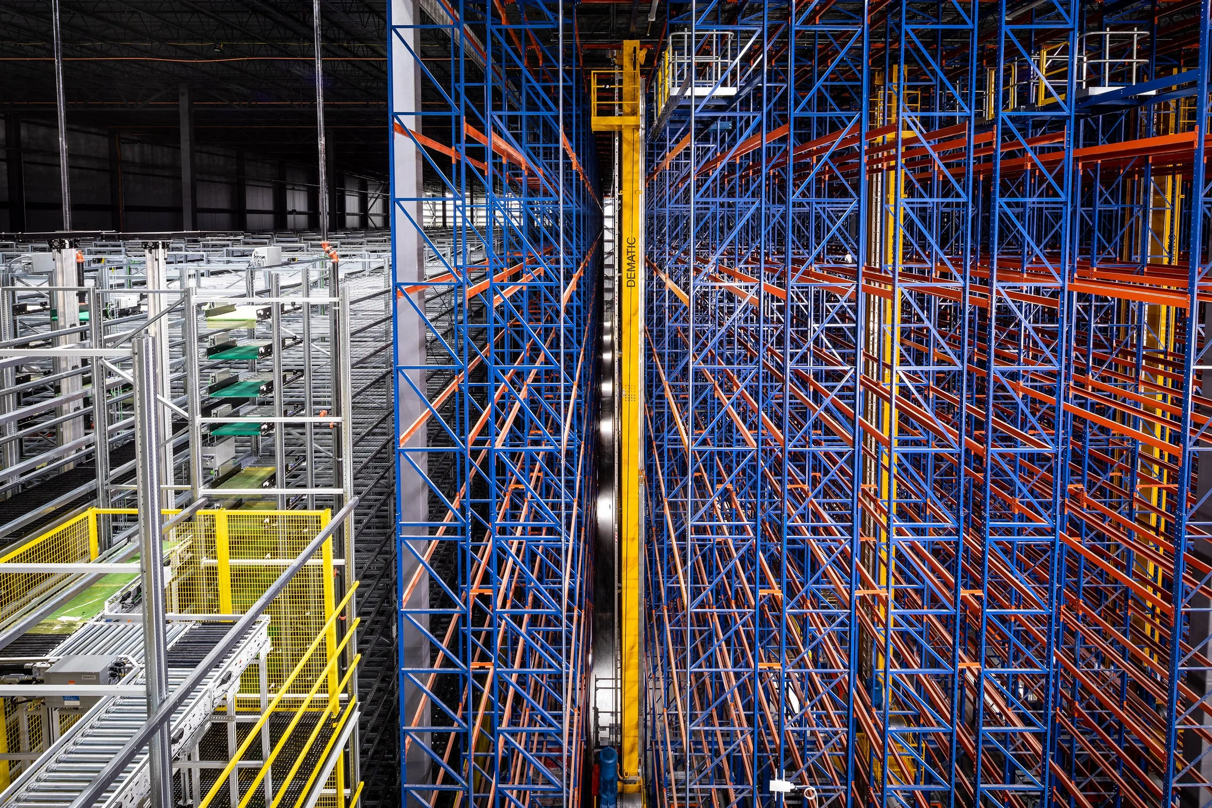 High-angle view inside a large distribution warehouse with tall, colorful metal storage racks and automated conveyor systems, illuminated by overhead lights.