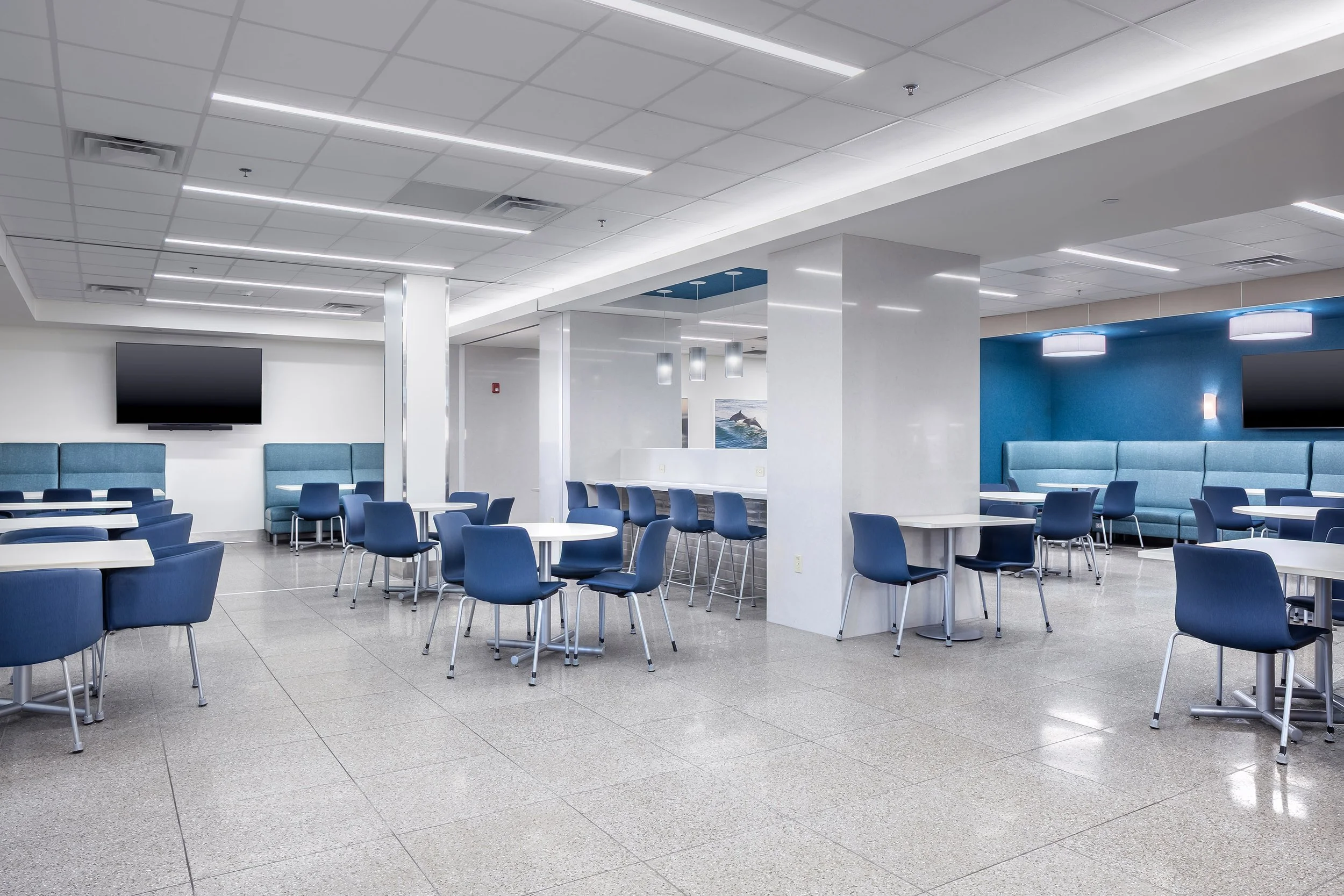 A modern, spacious cafeteria with blue chairs, white tables, tiled floor, and wall-mounted TVs. The room features bright lighting, cushioned booth seating, and clean interiors perfect for a healthcare setting.