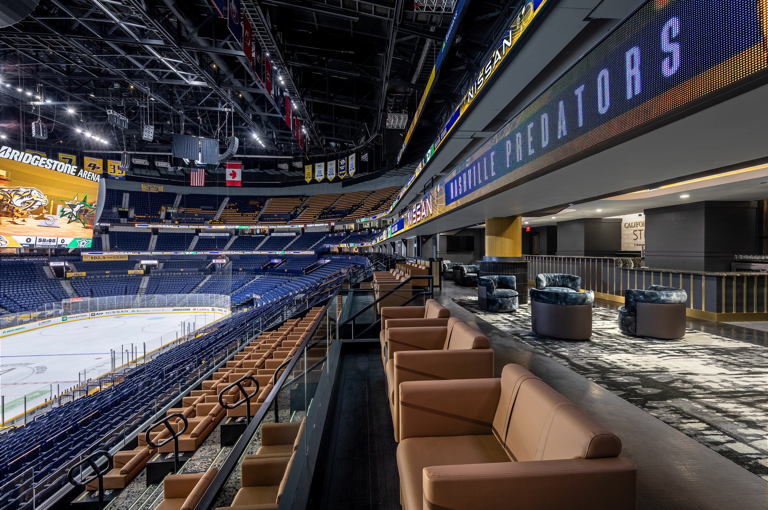 Luxury club suite seating area overlooking an empty ice hockey rink at Bridgestone Arena, with rows of tan chairs, lounge seating, and digital signs displaying “Nashville Predators.”.