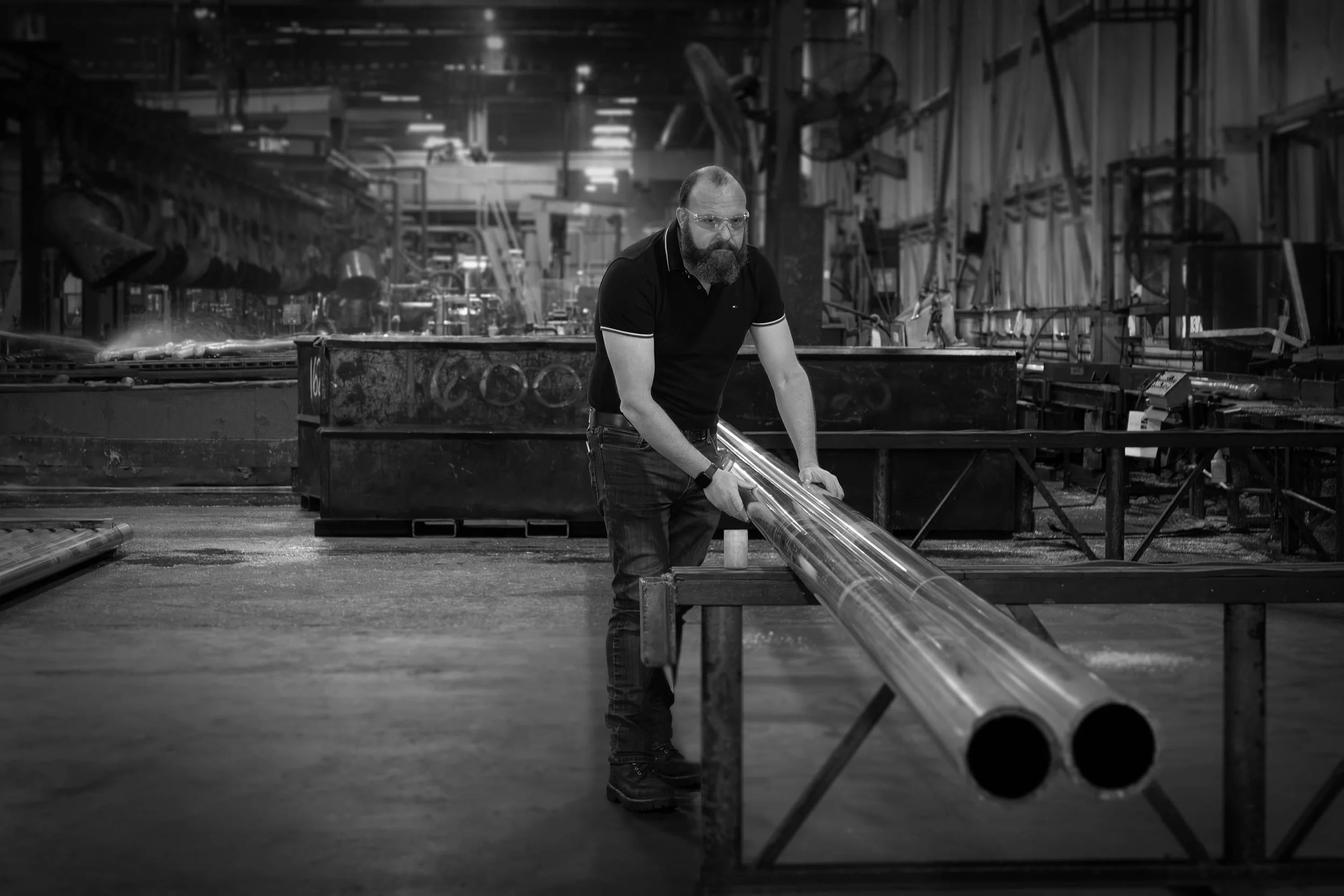A man in a factory moves large aluminum pipes on a workbench; the black and white scene highlights the dedicated workforce.