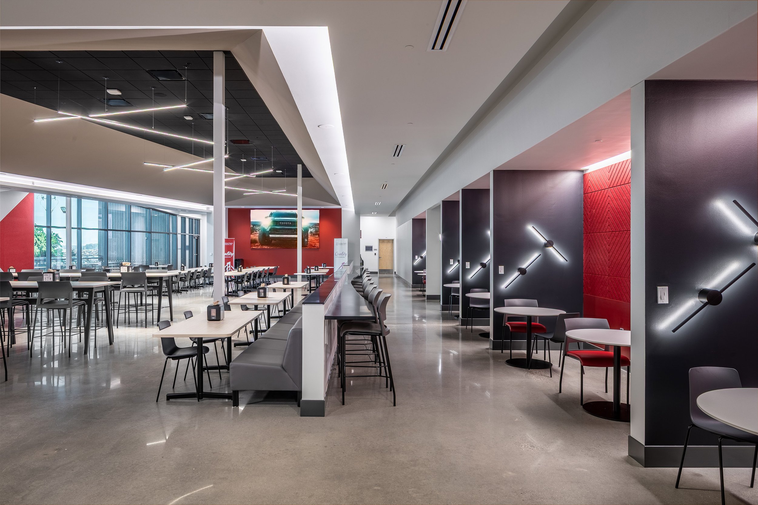 Modern cafeteria with long tables, high-top seating, and red accent walls. Geometric light fixtures decorate the walls, and large windows let in natural light. The space is clean and spacious with polished floors.