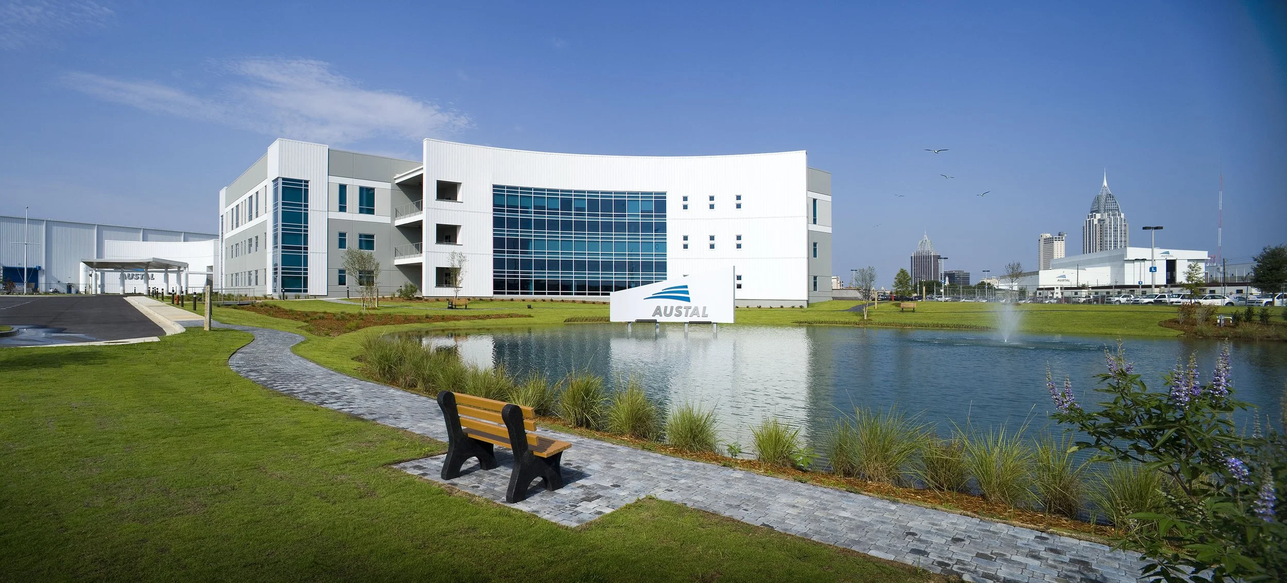 Modern office building beside a pond with a bench and walking path, featuring the Austal company logo.