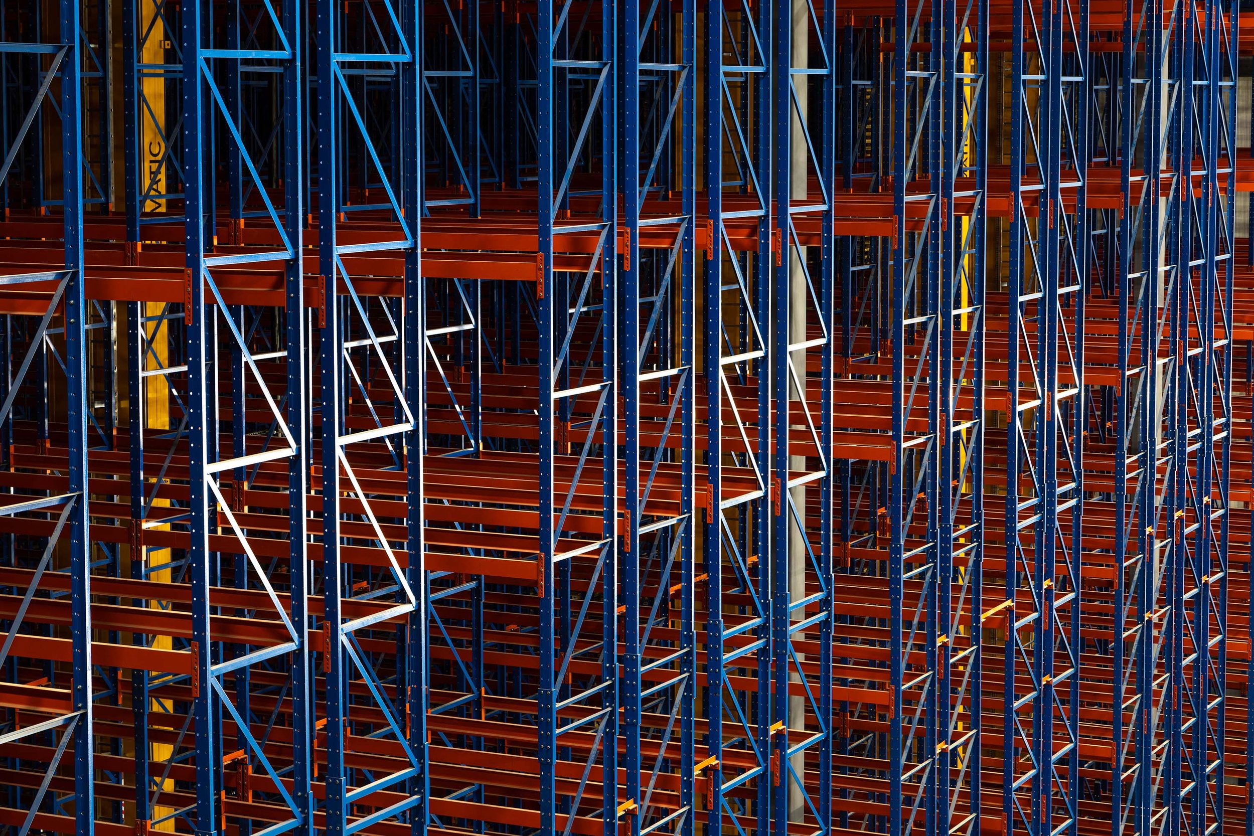 Rows of empty blue and orange racking shelves in a warehouse form a geometric pattern of intersecting lines and structures, with no products or items stored on them—used for automated distribution systems.