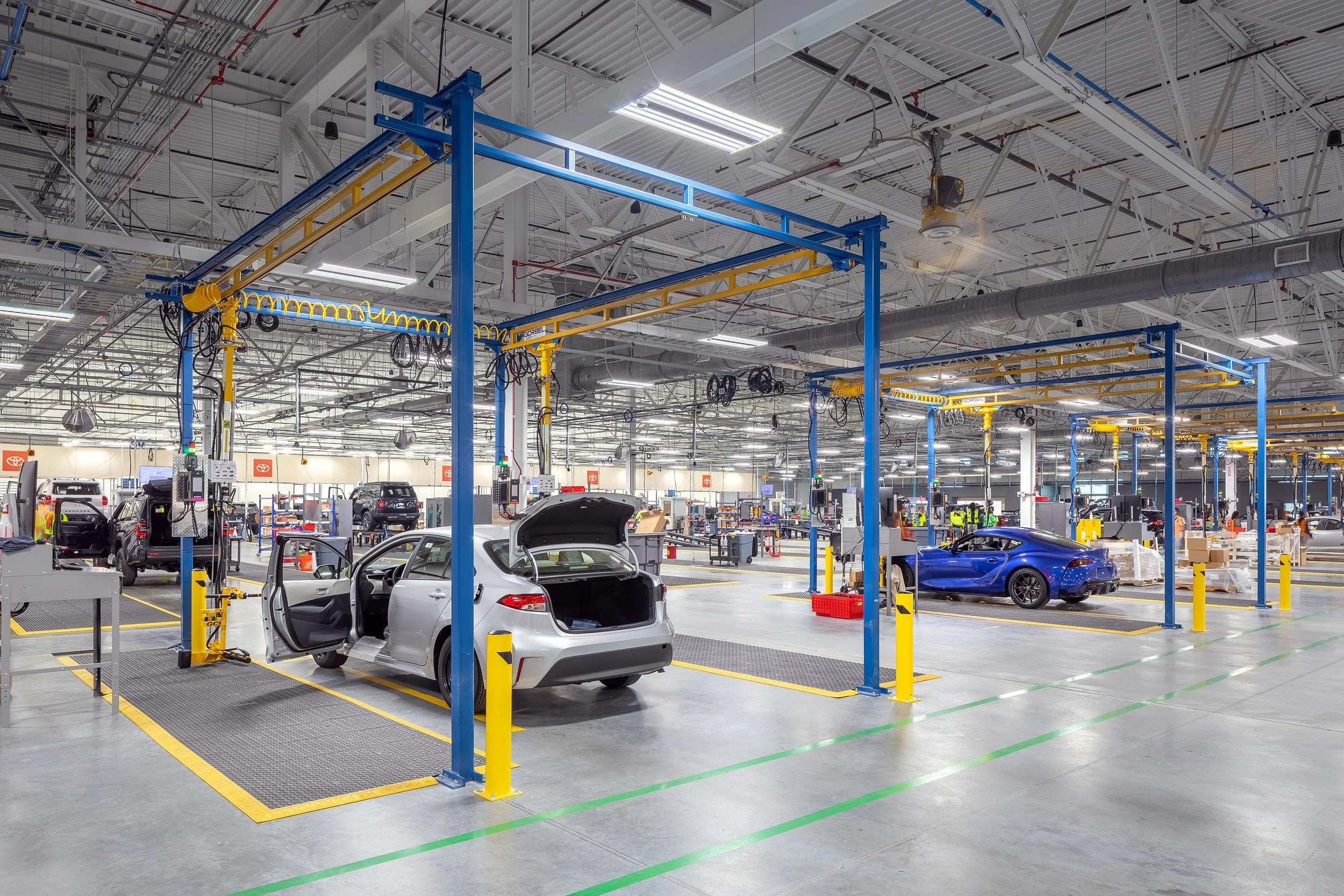 A spacious, brightly lit automotive factory interior with several cars being assembled. Open car doors and hoods are visible, and workers are seen in the background among workstations and equipment.