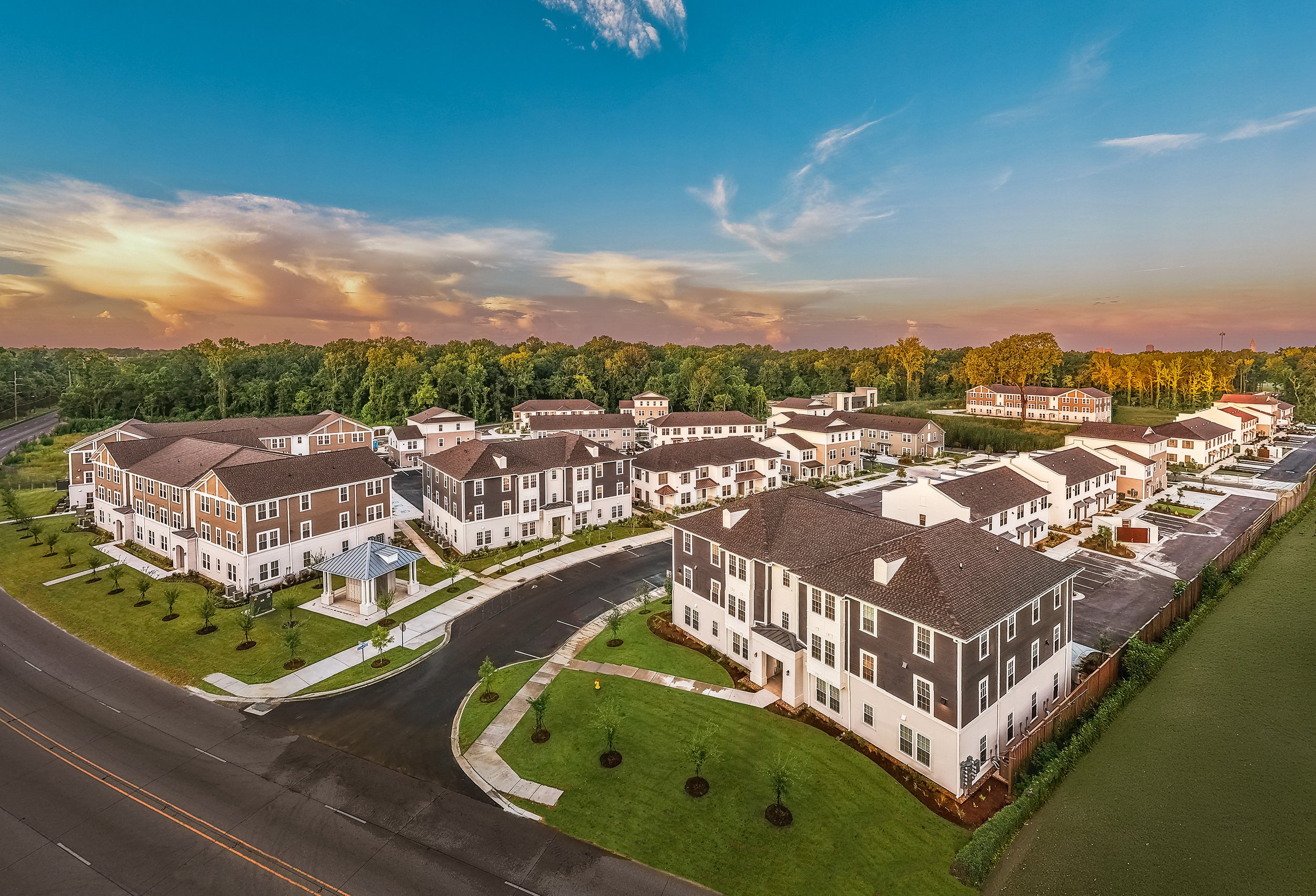 Aerial view of a modern residential complex with multiple large, three-story buildings, green lawns, trees, and a clear sky at sunset. Walking paths and driveways curve between the buildings.