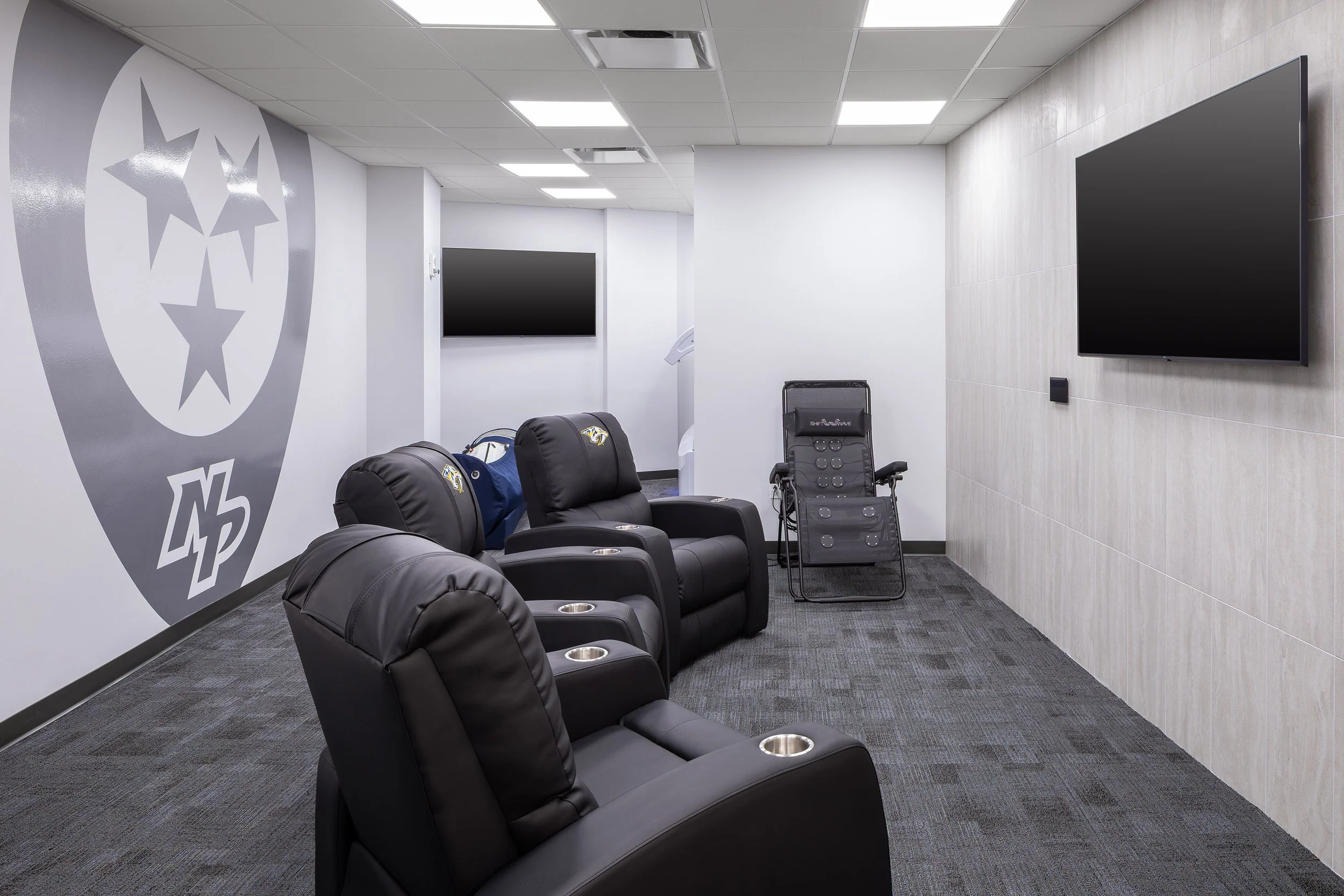 A modern lounge with three black recliner chairs facing wall-mounted flat-screen TVs, sports-themed wall art featuring hockey and arena vibes, and a massage chair in the back corner on a gray carpeted floor.