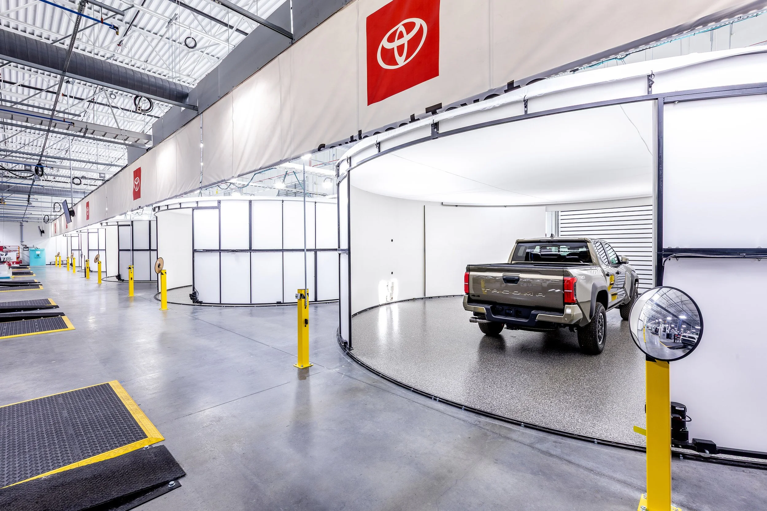 A silver Toyota Tacoma pickup truck is parked inside a bright, circular inspection bay at a modern automotive service facility, with Toyota logos visible above several adjacent workstations.