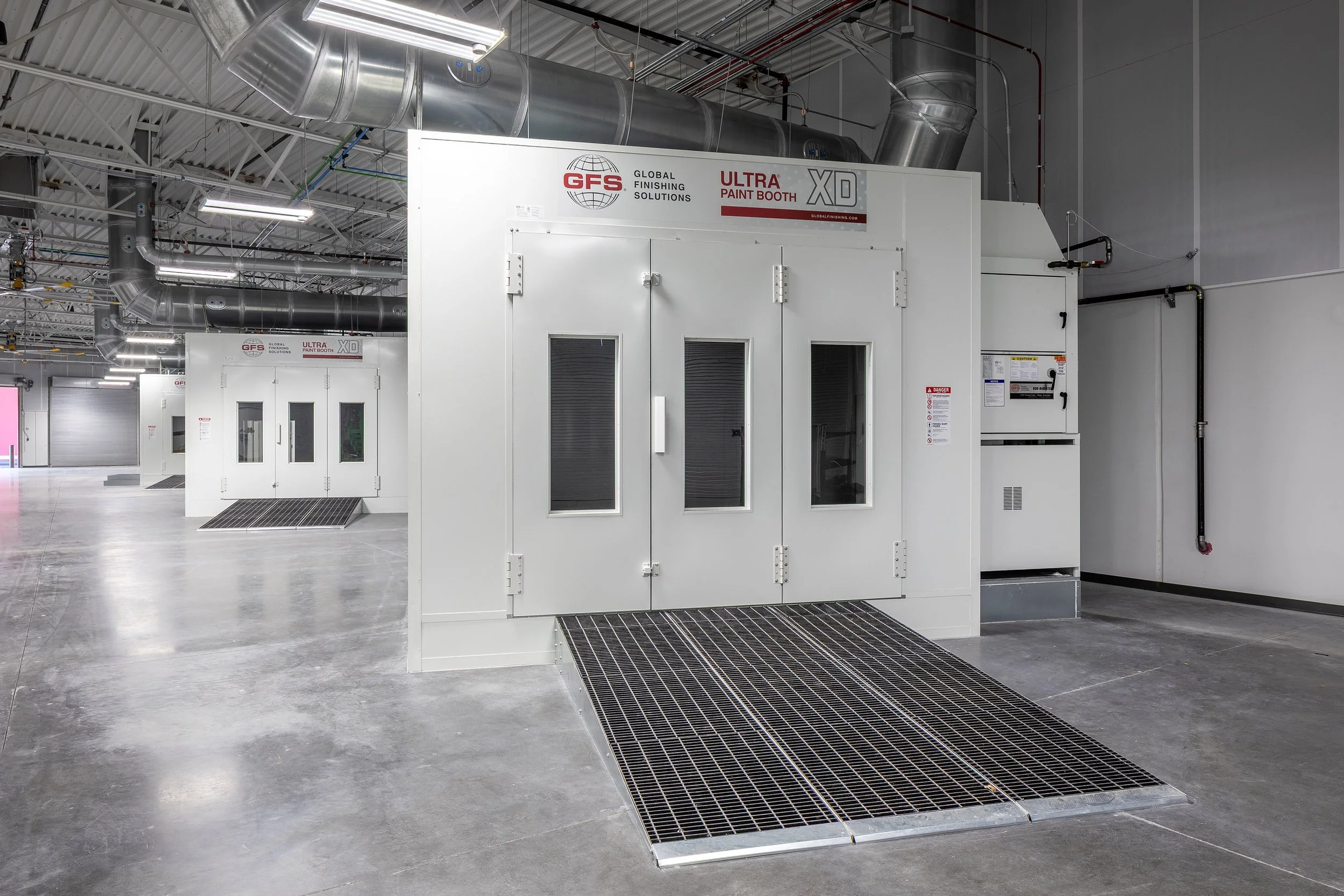 A modern industrial paint booth with large doors, ventilation grates, and overhead ductwork in a spacious, clean facility with polished concrete floors and bright lighting.