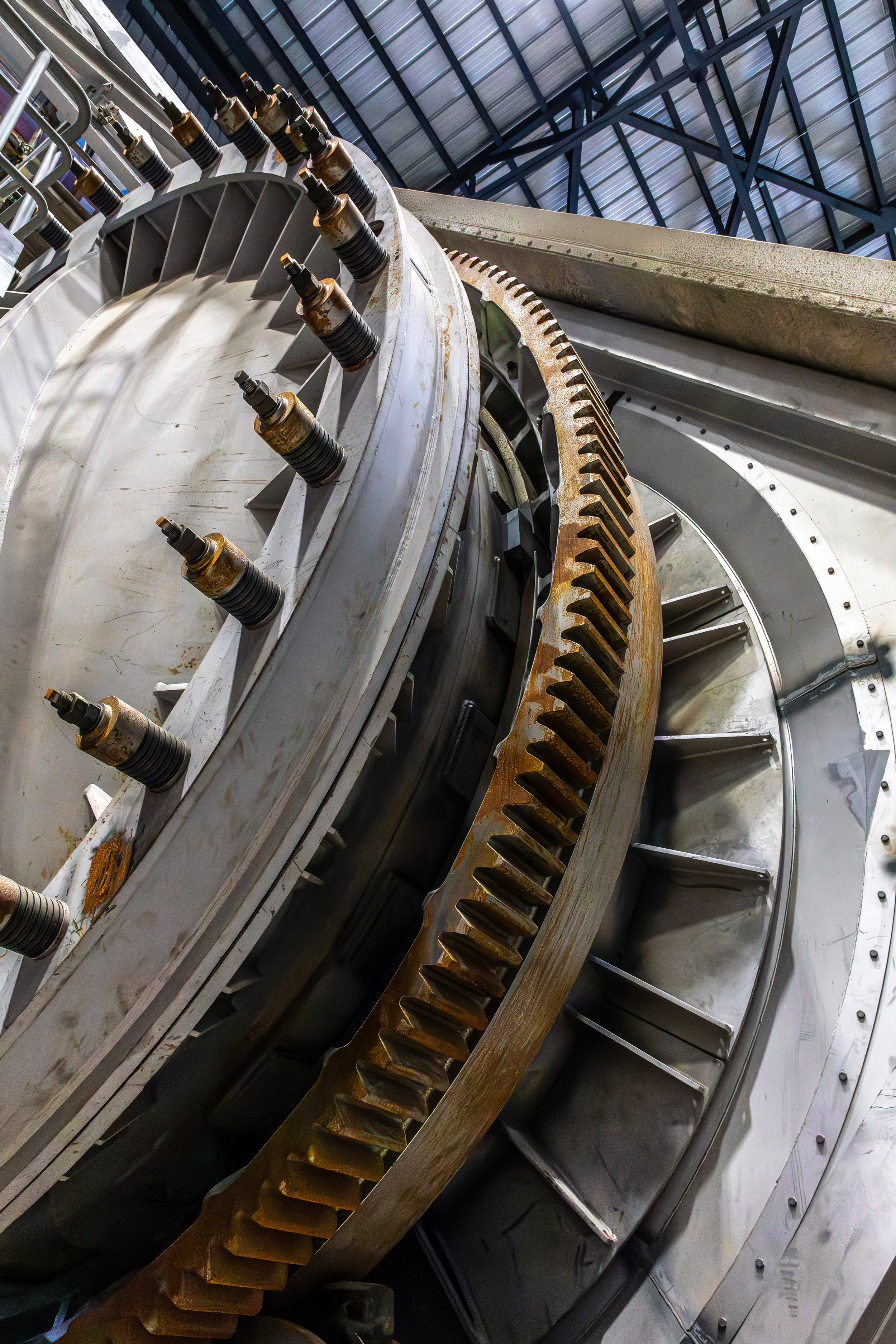 Close-up photography of large industrial metal gears and bolts inside a factory, with a high ceiling and structural beams visible in the background.