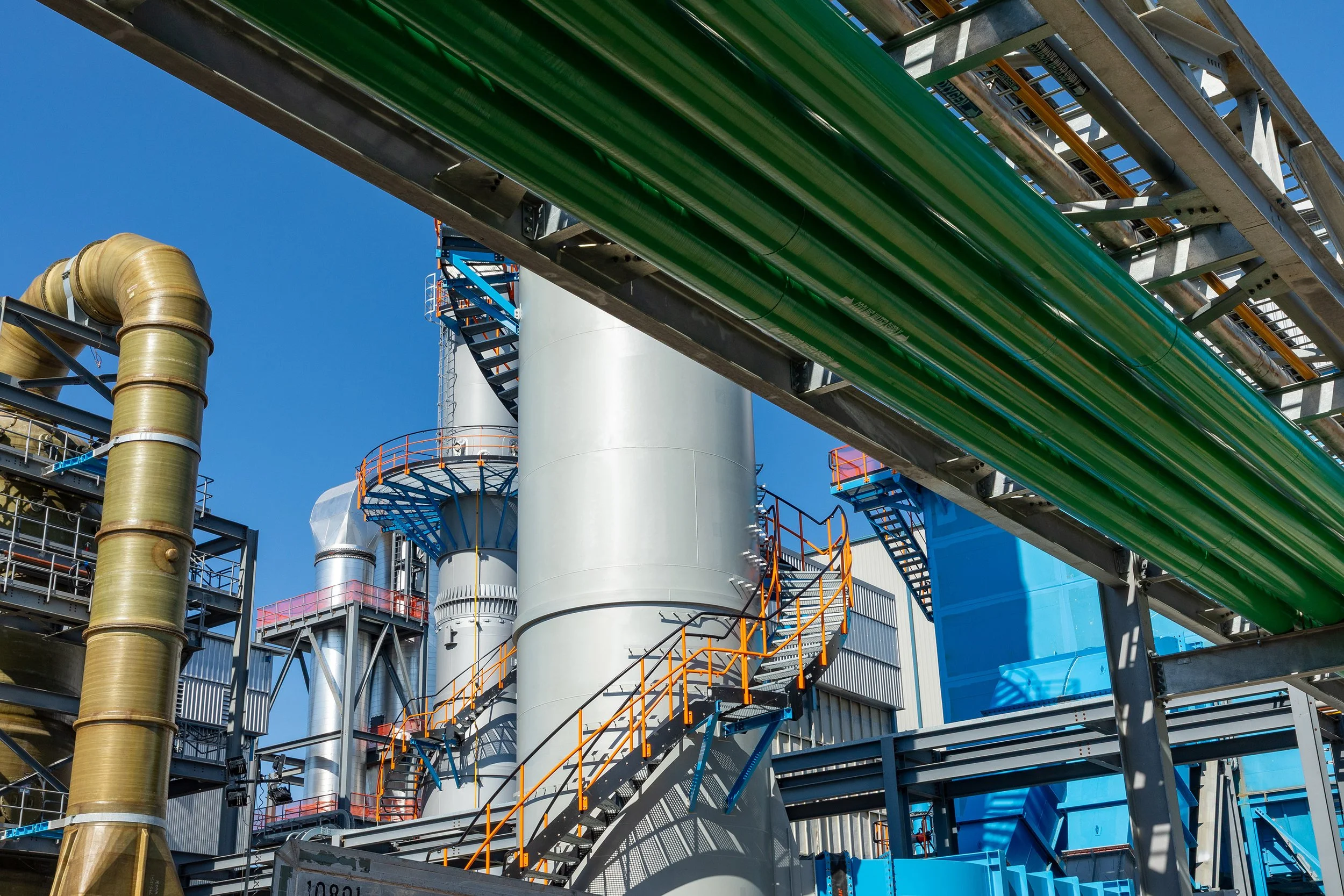 Industrial facility with large metal tanks, green pipes, staircases, and platforms against a clear blue sky. This industrial scene captures complex machinery and metals structures, typical of a factory or processing plant.
