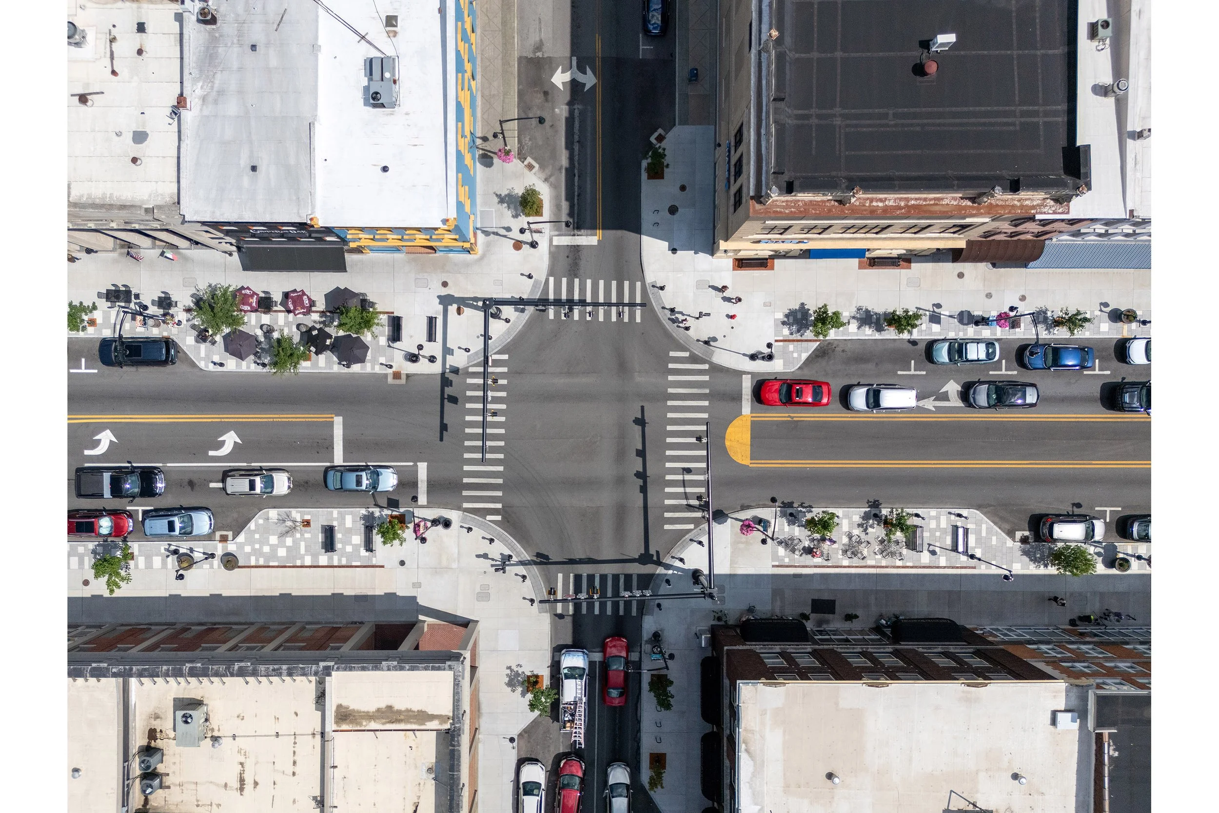Aerial view of a main street intersection with crosswalks, cars parked along the streetscape, and buildings on each corner.