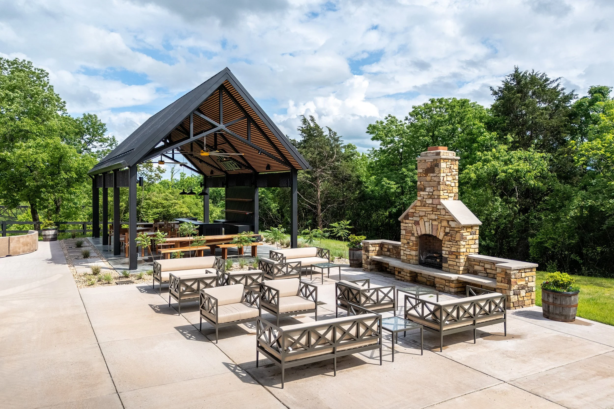 Outdoor patio with metal chairs arranged around a stone fireplace. A pavilion with a peaked roof covers picnic tables. Green trees and a partly cloudy sky set the scene—perfect for photography or sipping bourbon in a relaxing atmosphere.