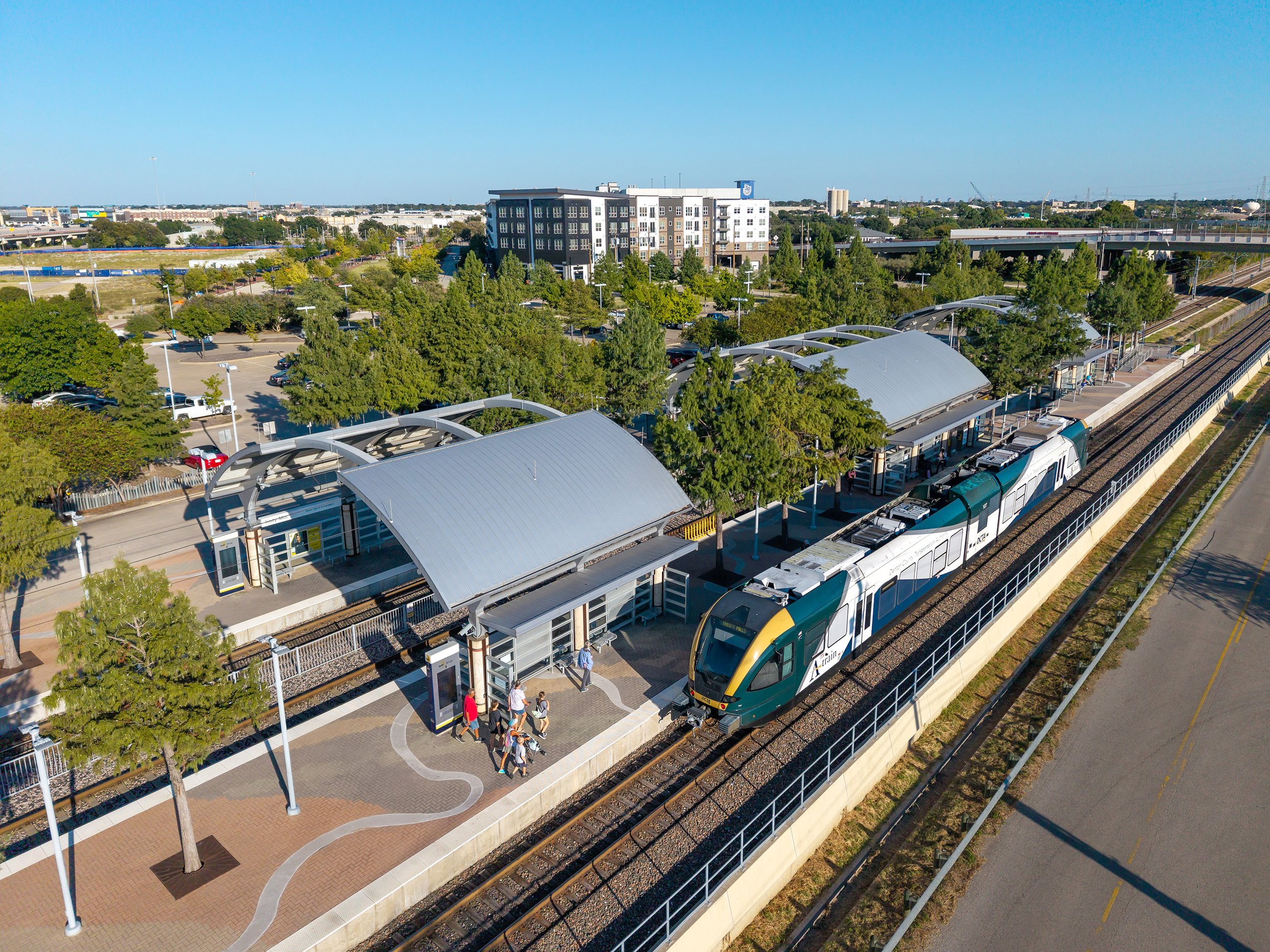 A modern light rail train stops at an outdoor station with curved metal canopies, surrounded by trees and people boarding, with buildings and a parking lot in the background under a clear blue sky.