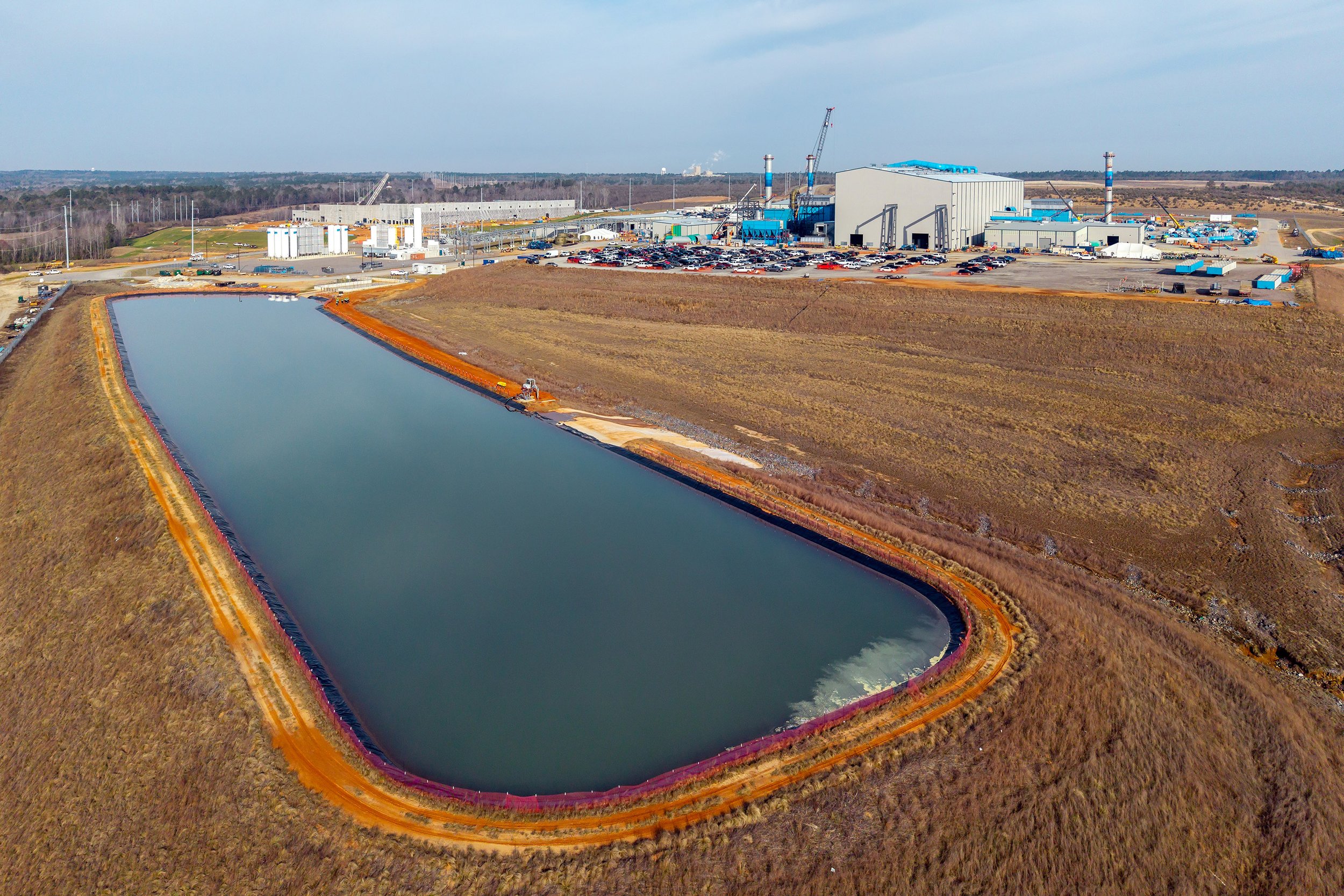 A large industrial facility sits near a wide, rectangular retention pond surrounded by grasses and red soil, where metals processing equipment and construction vehicles are visible in the background.