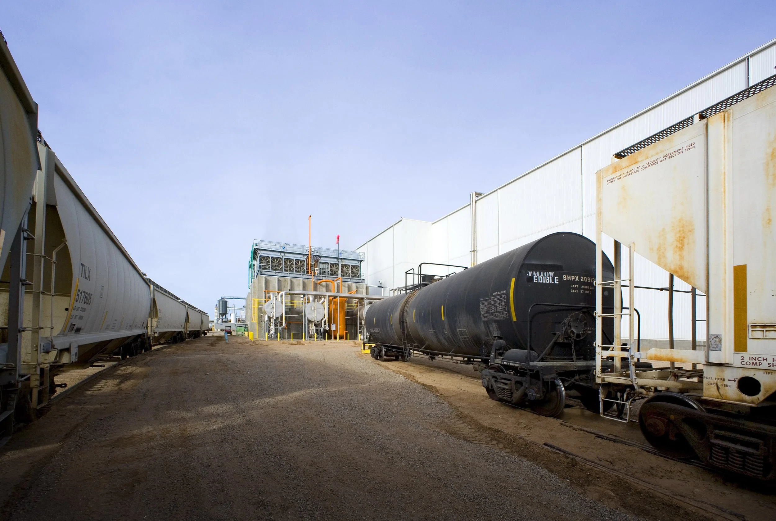 Industrial food facility with large white and black rail cars on gravel between tall white buildings under a clear sky.