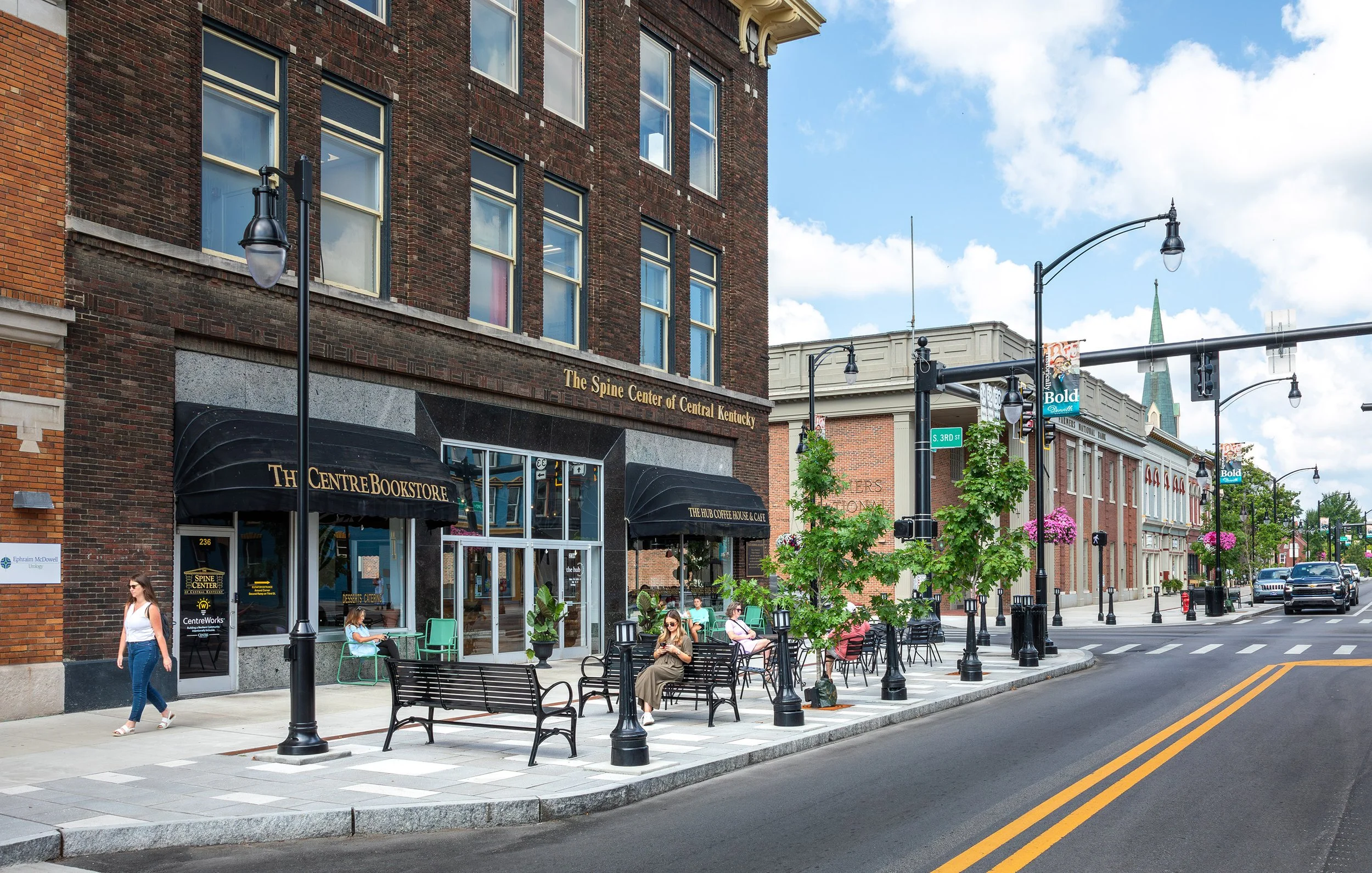 People sit on benches outside a historic bookstore, surrounded by shops and trees on a city street under a partly cloudy sky.