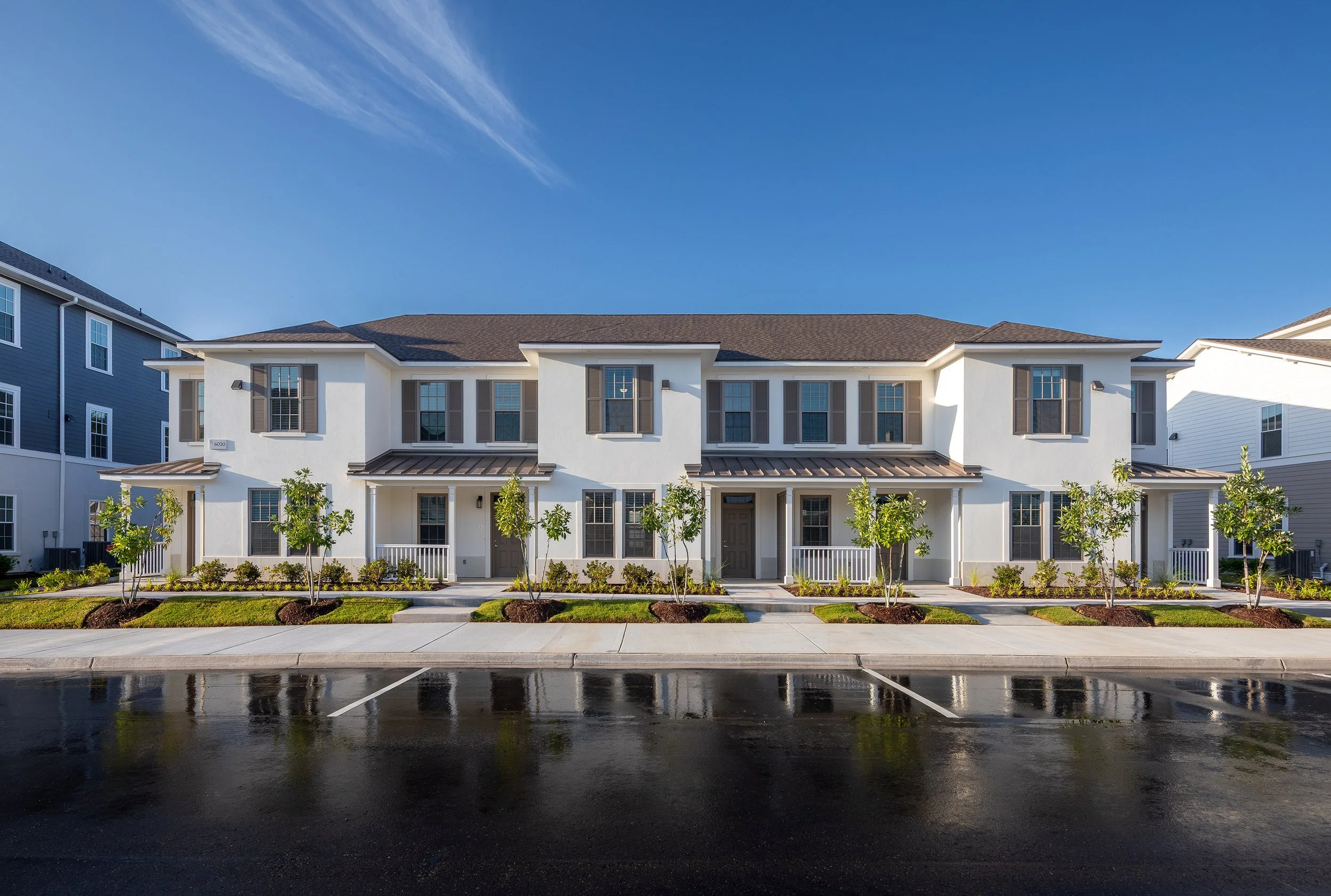 A modern two-story townhouse building with white walls, gray accents, multiple windows, and small front porches. Young trees and neatly trimmed bushes line the sidewalk, with a parking lot and clear blue sky in front.