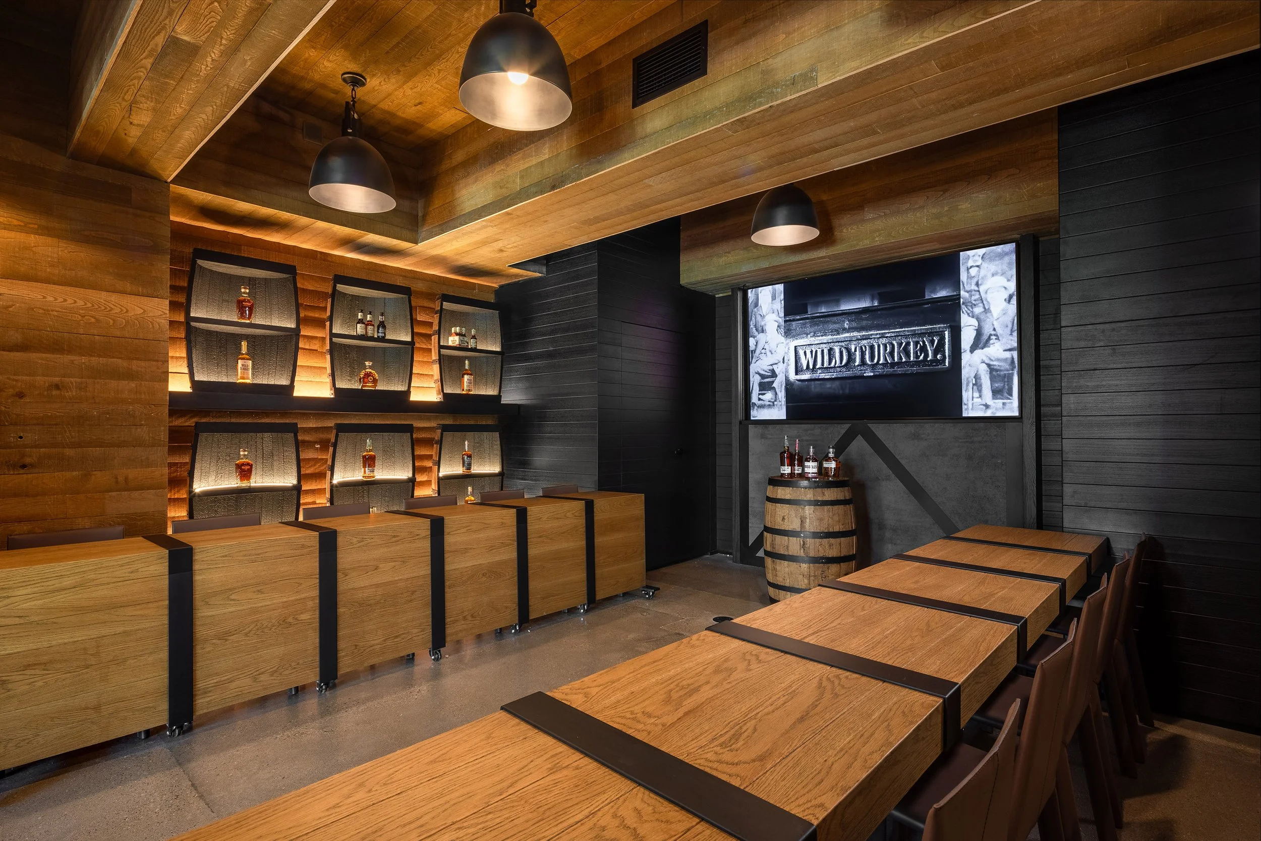 A modern tasting room with wooden walls and ceilings, a display of bourbon whiskey bottles, a long table with chairs, a barrel with bottles, and a large screen showing Wild Turkey at the visitor center.