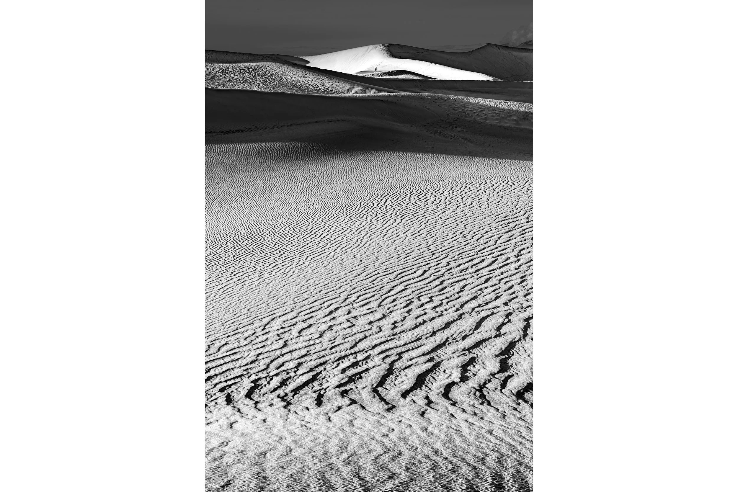 Black and white photo of Death Valley sand dunes with rippled patterns and a bright, smooth dune in the background.