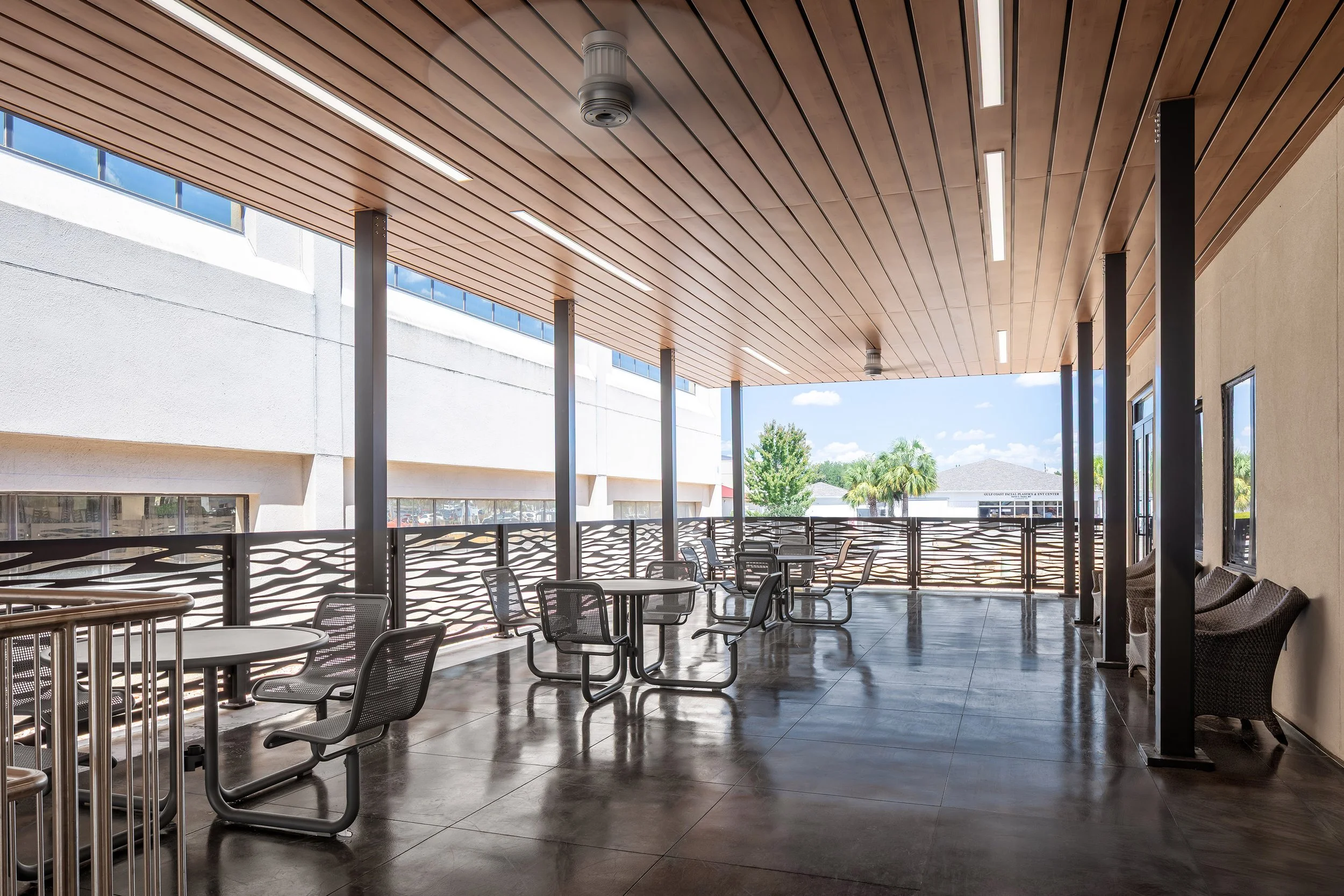 A covered outdoor patio with modern metal tables and chairs, ceiling fans, and a decorative railing. Large windows on one side showcase sunny views of trees and mountains, perfect for photography enthusiasts who love scenic interiors.